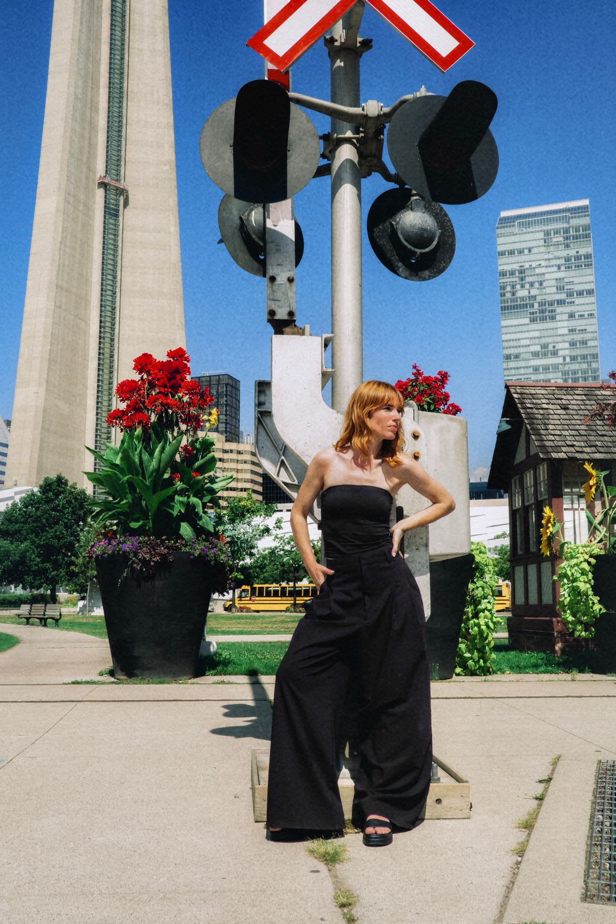 A woman in a black strapless outfit poses confidently by a railroad crossing signal in a city park on a sunny day.