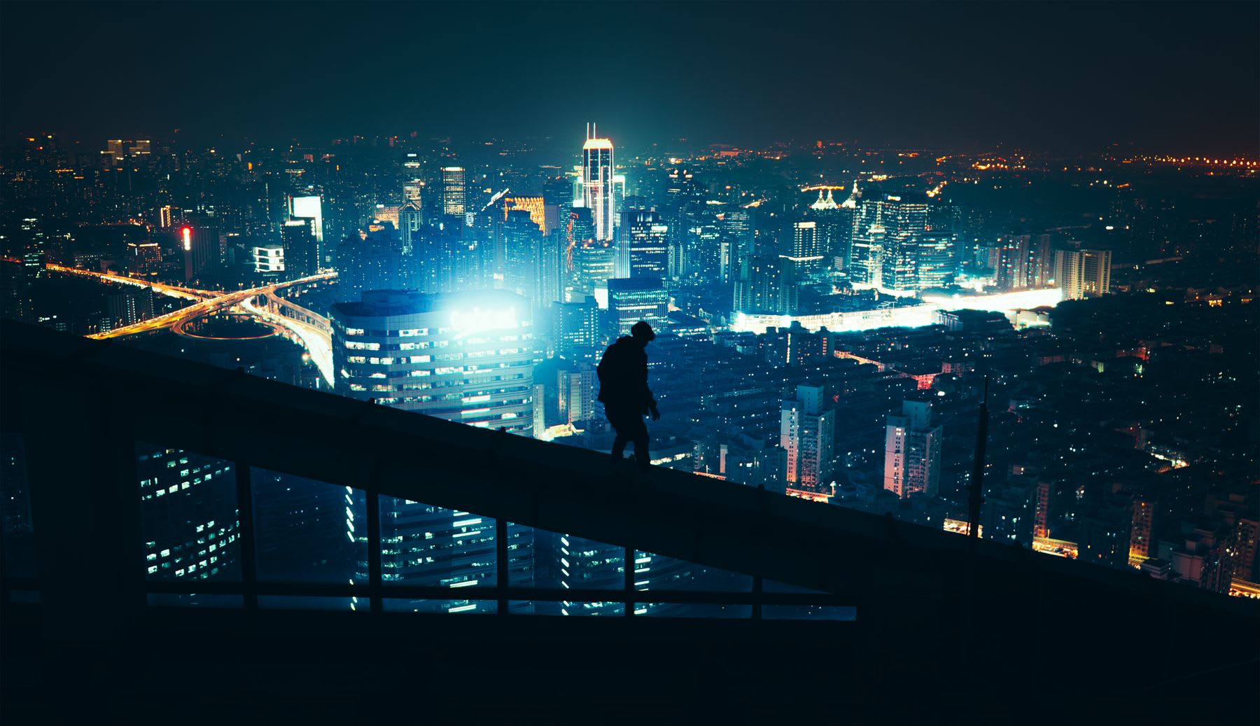 A man walking at the edge of a skyscraper's roof at night, with the city skyline illuminated by numerous lights in the background.