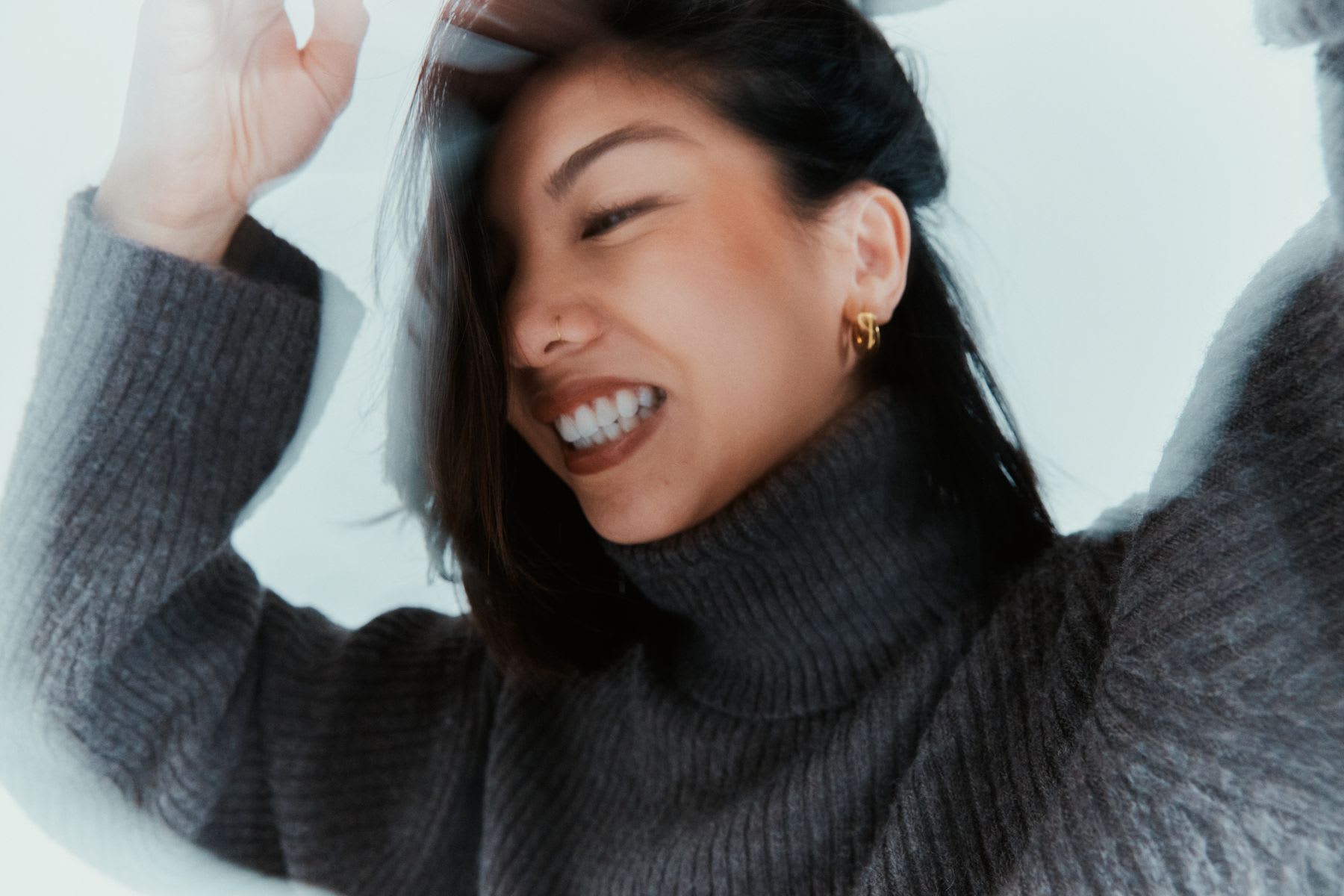 Smiling woman with a golden earring wearing a gray turtleneck and with her hands raised.