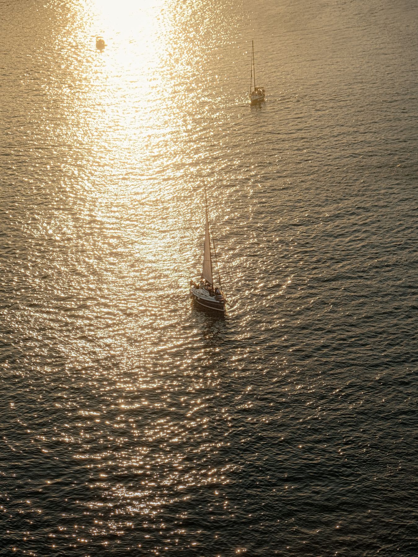 Sailboats navigate the open sea, their path illuminated by the brilliant golden reflection of the sun on the water.