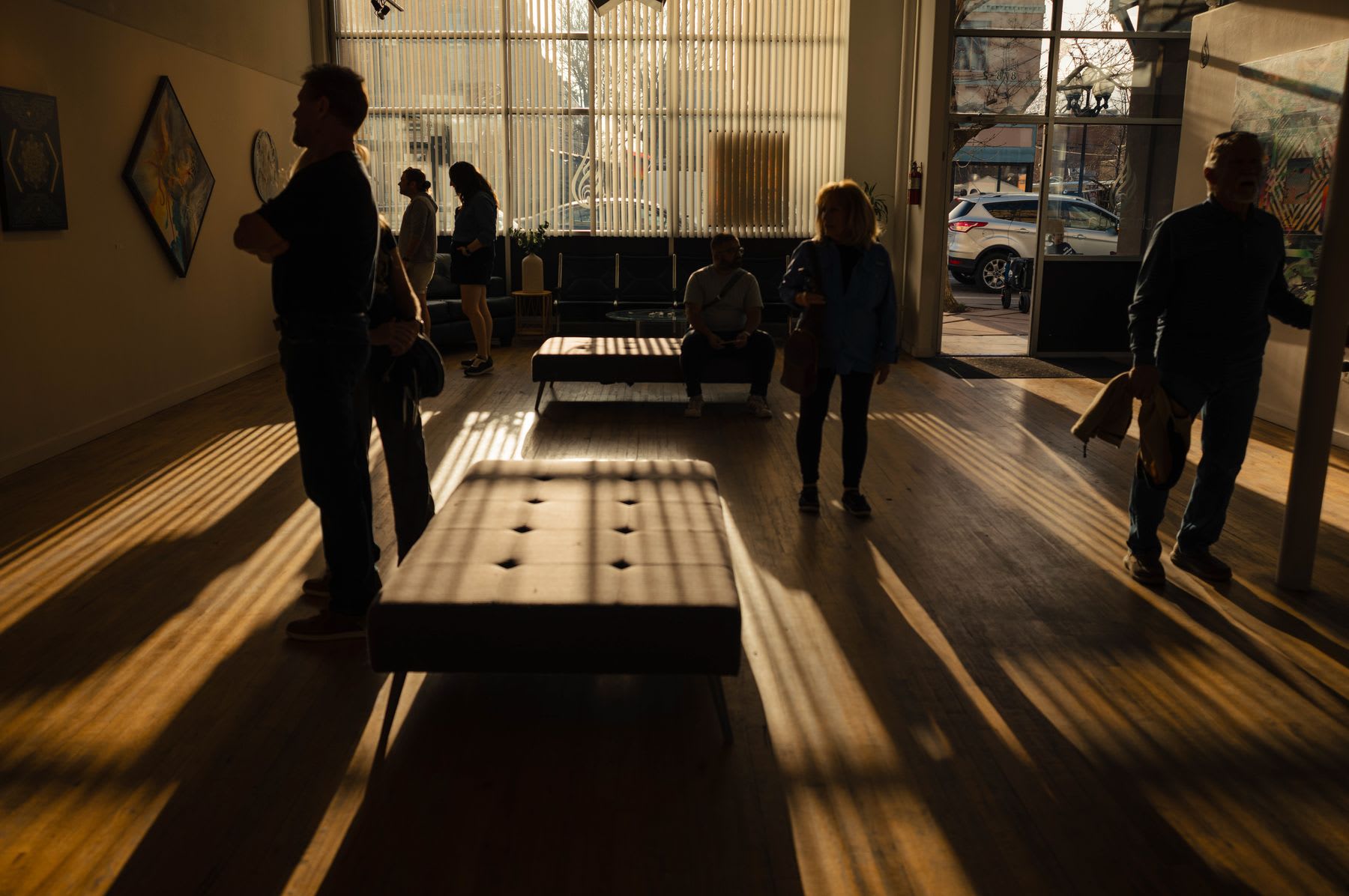 Several people are viewing art in a gallery with sunlight streaming in, casting long shadows on the wooden floor.