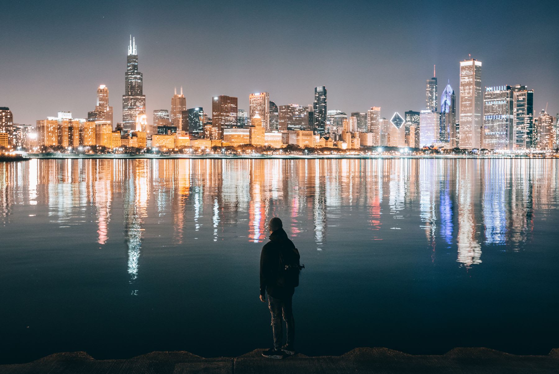 Man standing along the coast looking at the metropolitan skyline as city lights reflect upon the water