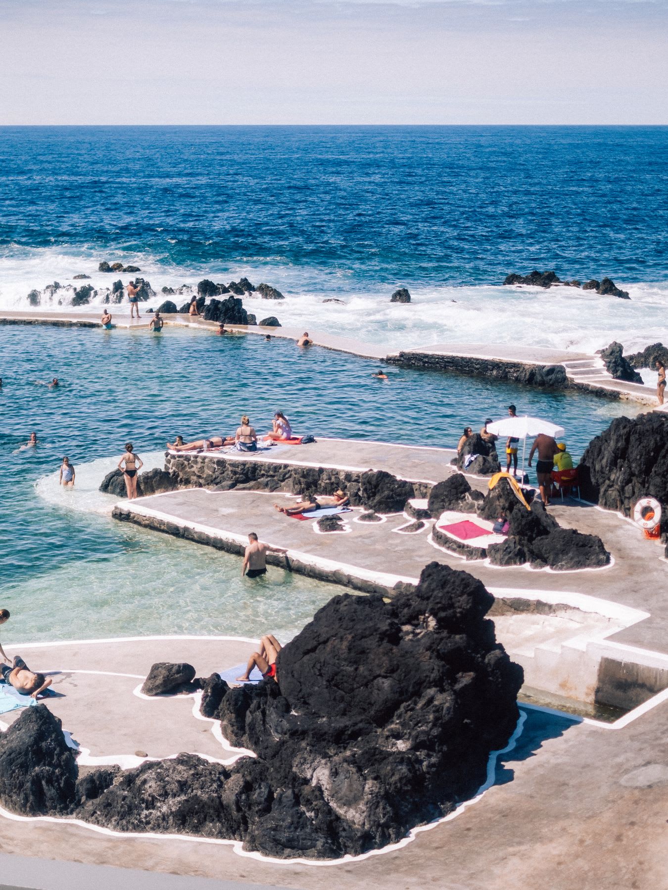 Natural ocean pool surrounded by black volcanic rocks, with people swimming and relaxing around.