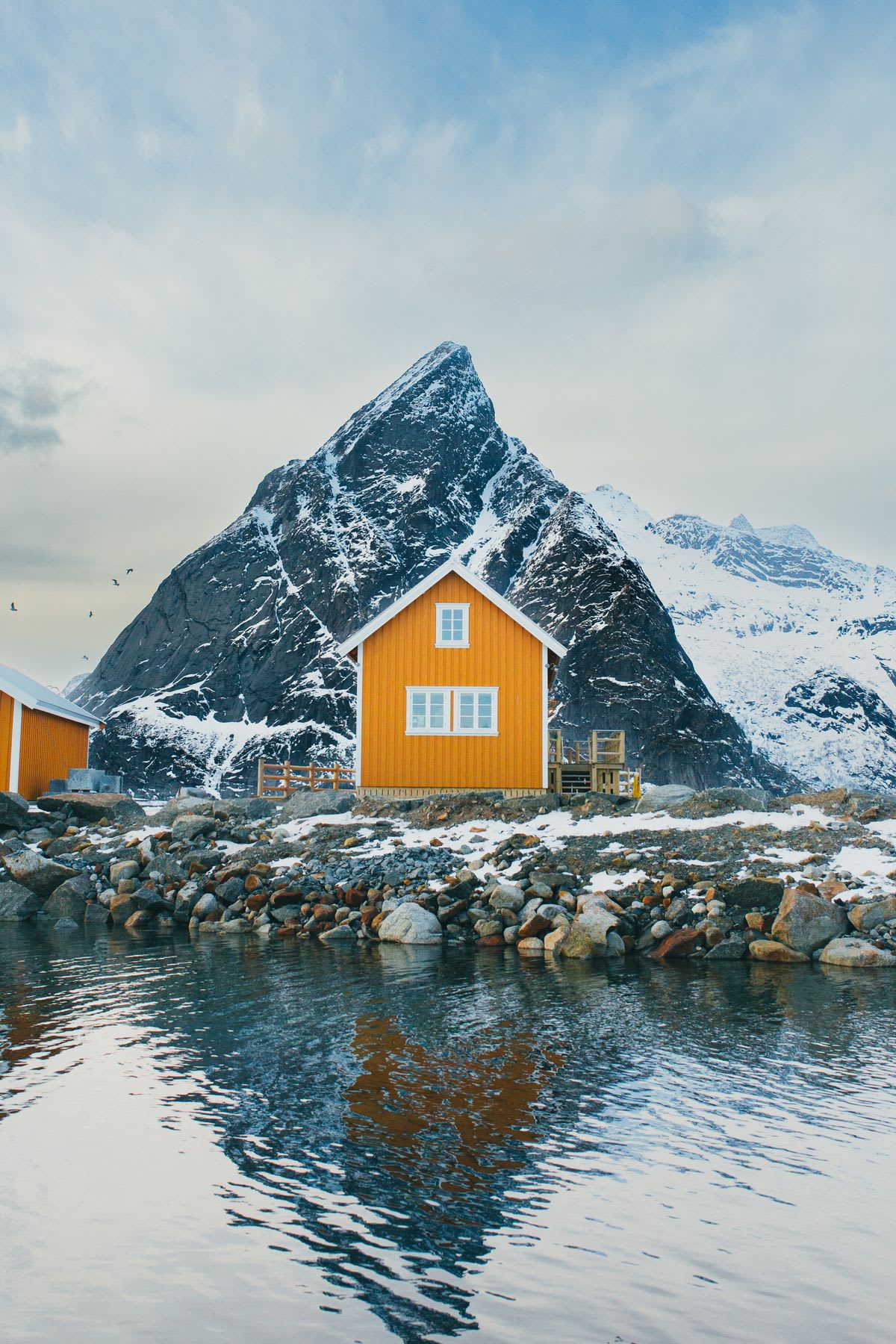 A yellow house in front of a snowy mountain, next to a body of water.