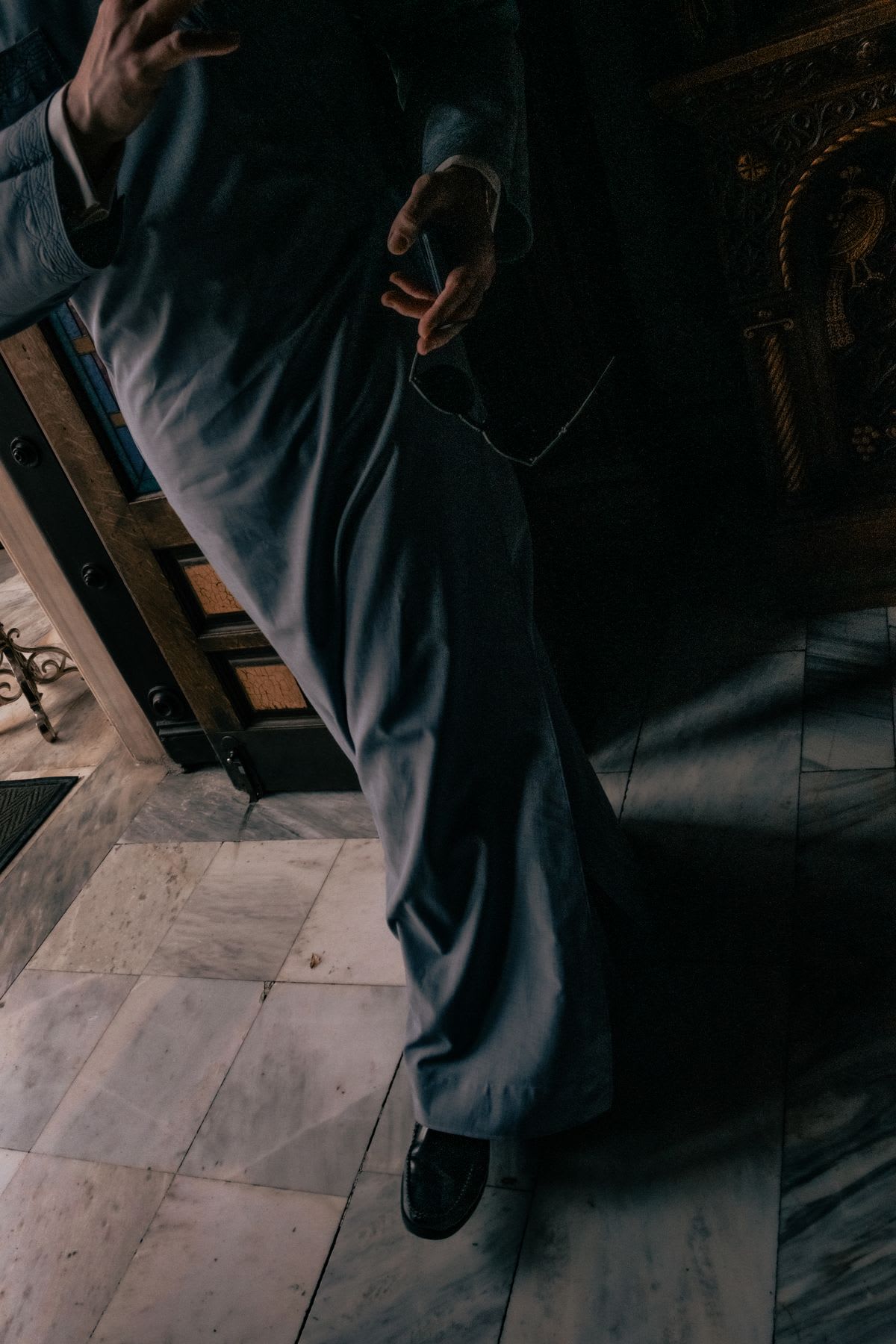 A low-angle view shows a man in a flowing grey garment walking across a marble floor, his hand holding a pair of glasses.