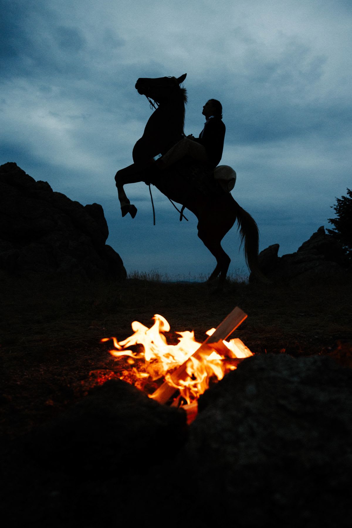 In a dimly lit twilight scene, the silhouette of a man on a rearing horse appears on rocky terrain, positioned behind a brightly burning campfire in the foreground.