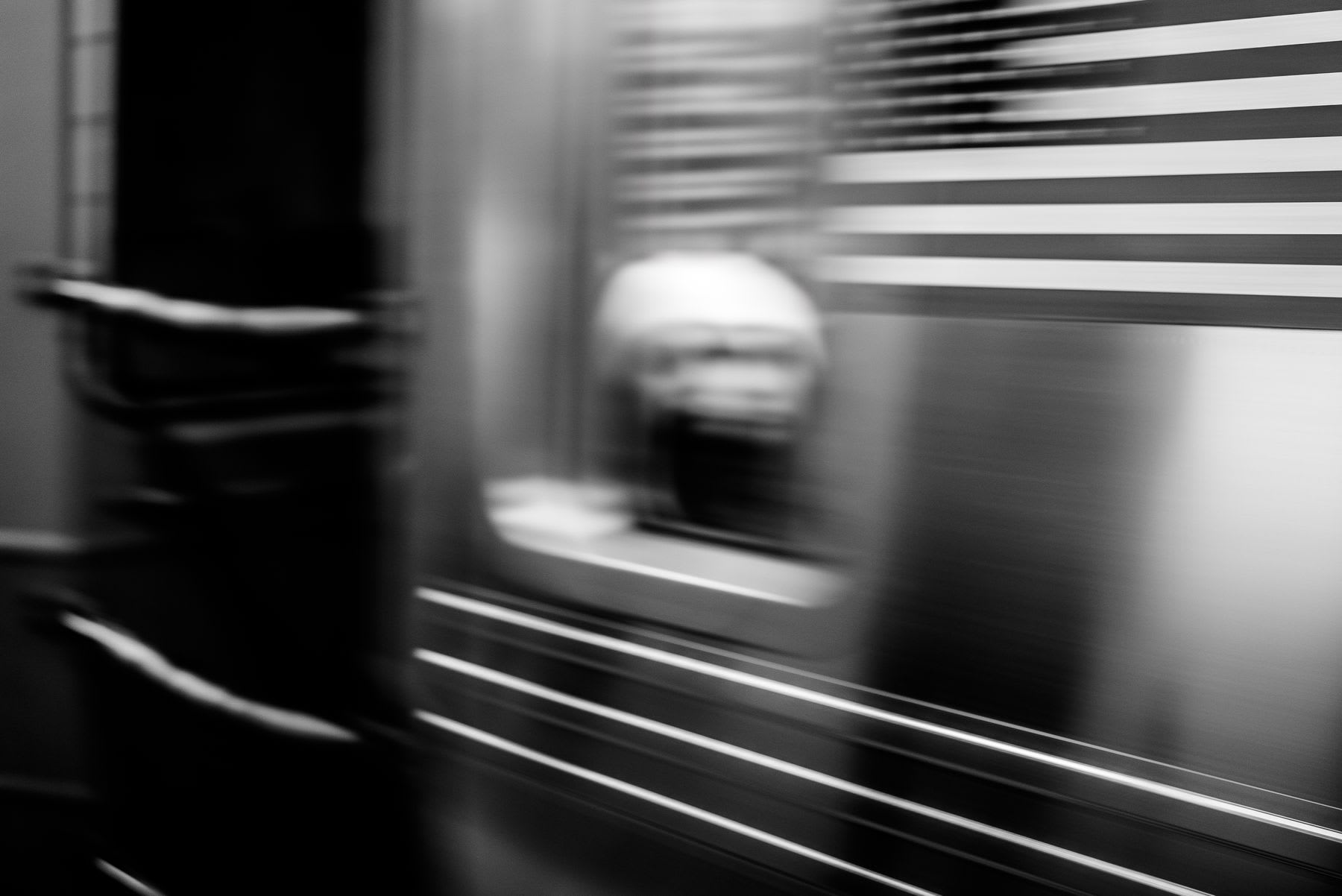 A motion-blurred monochrome shot glimpses a person in a speeding NYC subway car, with a striped, flag-like shape also blurred in the rush of movement.