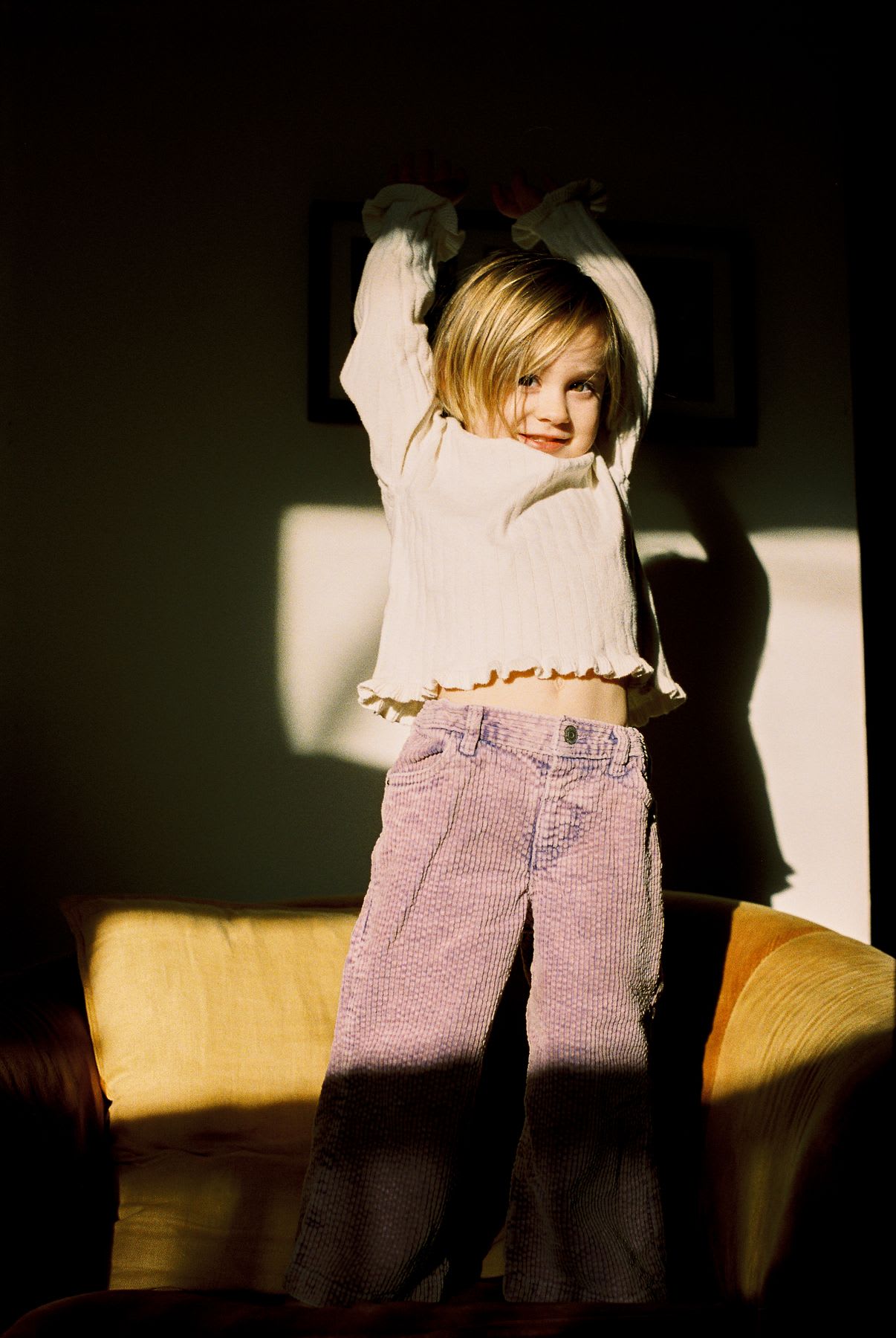 A young girl in a sunbeam playfully stretches her arms while standing on a yellow couch in a dark room.