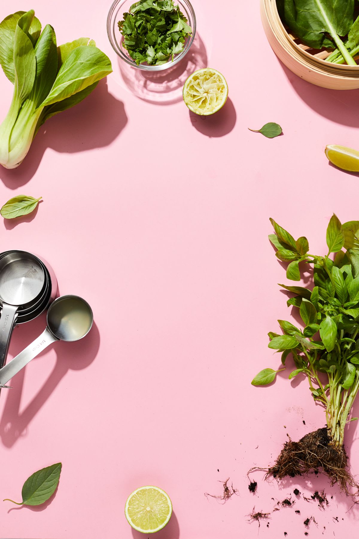 Fresh herbs, lime halves, and bok choy are artfully arranged on a pastel pink surface alongside metal measuring spoons