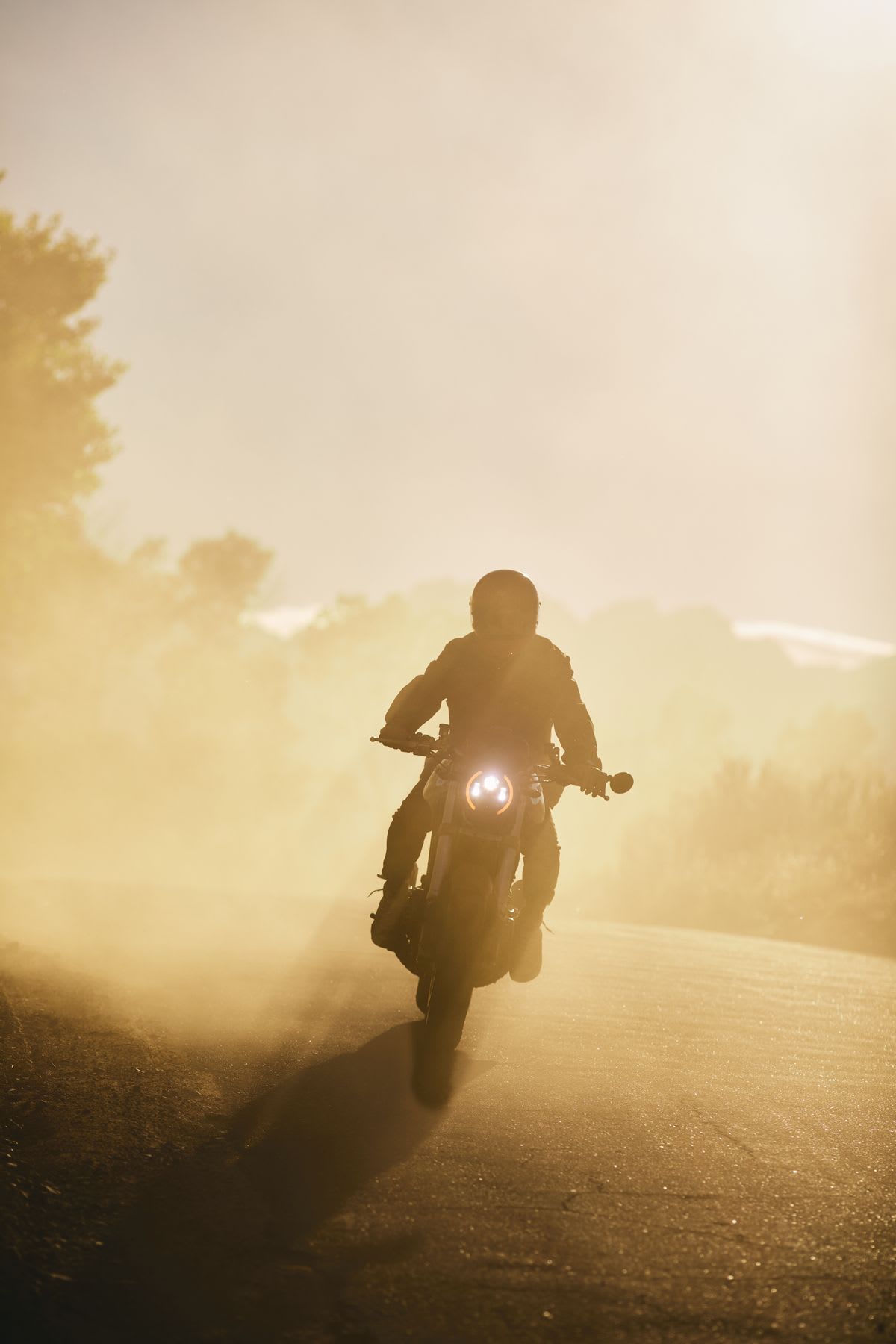 A lone motorcyclist kicks up a thick cloud of dust while riding down a rural dirt road during the golden hour, creating a dramatic silhouette against the bright, hazy sunlight with the motorcycle's single headlight piercing through the gloom.