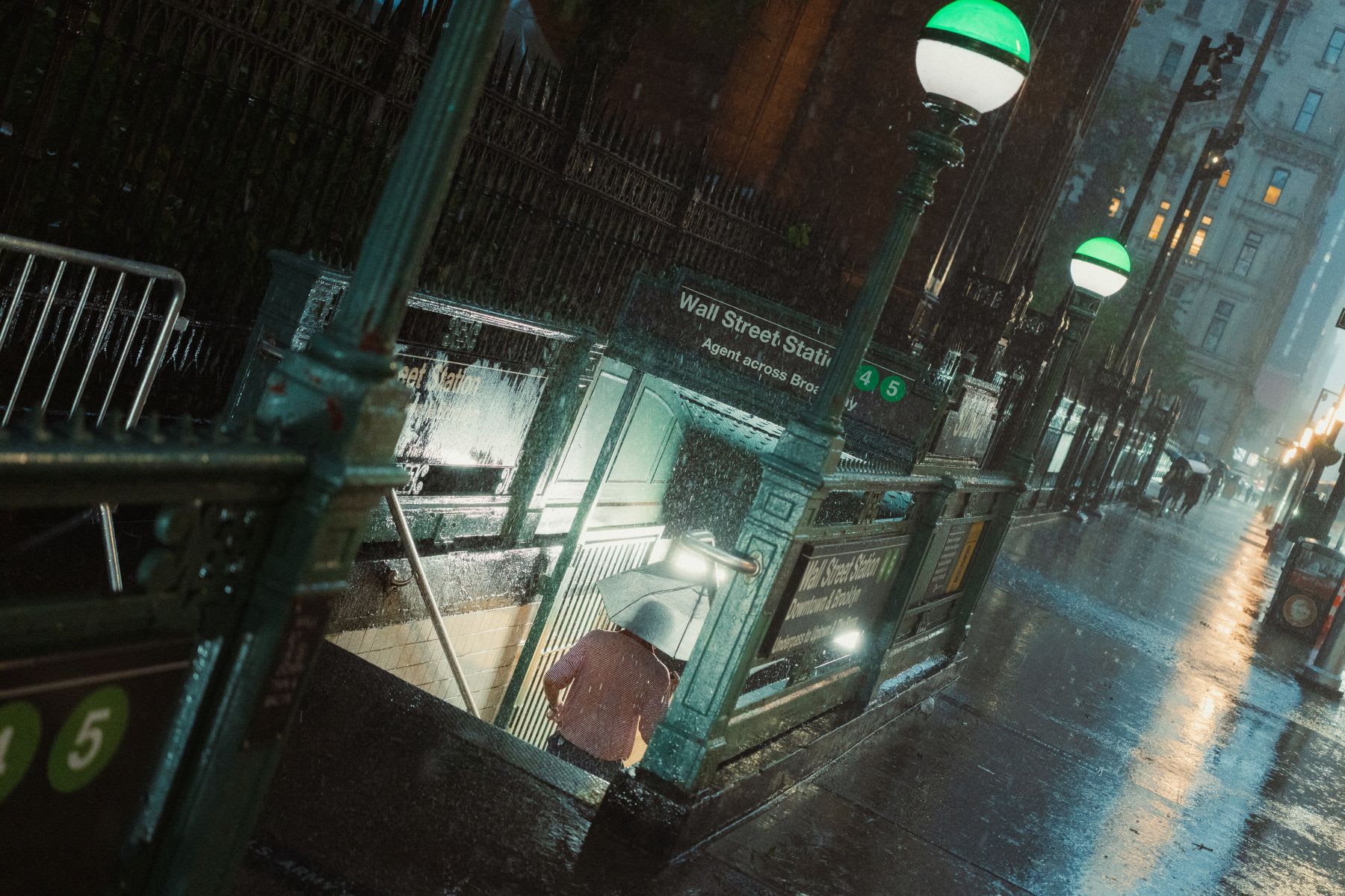 A person with an umbrella descends into the Wall Street subway station during a heavy downpour on a city street at night.