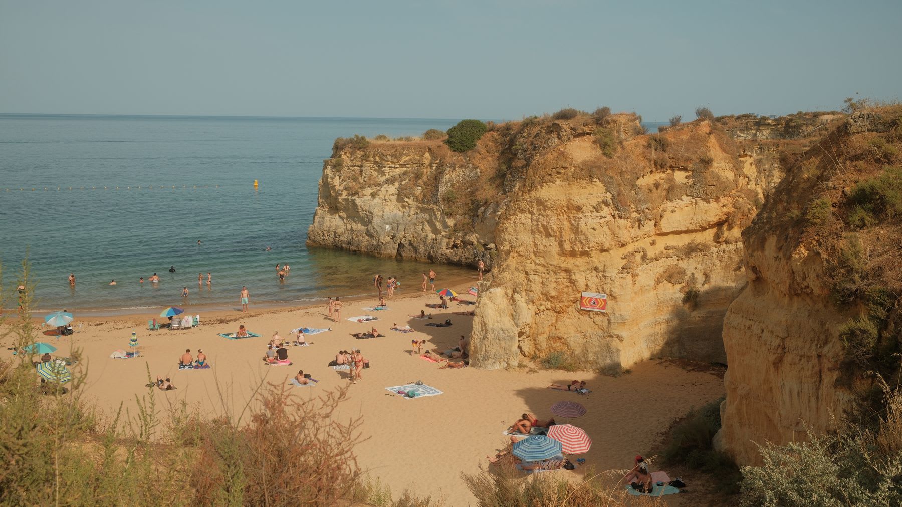 A high-angle view shows people enjoying a sunny day on a sandy beach nestled between golden cliffs and the calm sea.