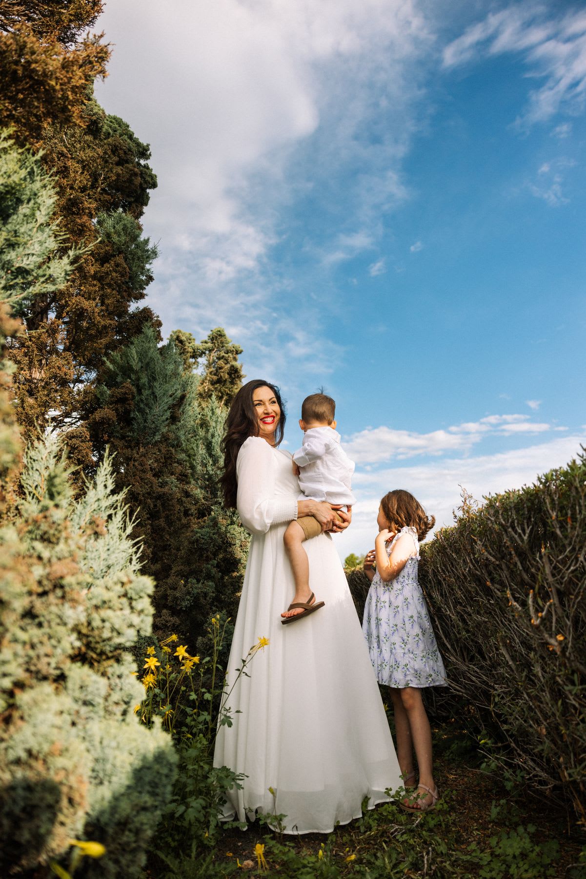 A woman in a white dress holding a young boy and standing next to a young girl in nature.