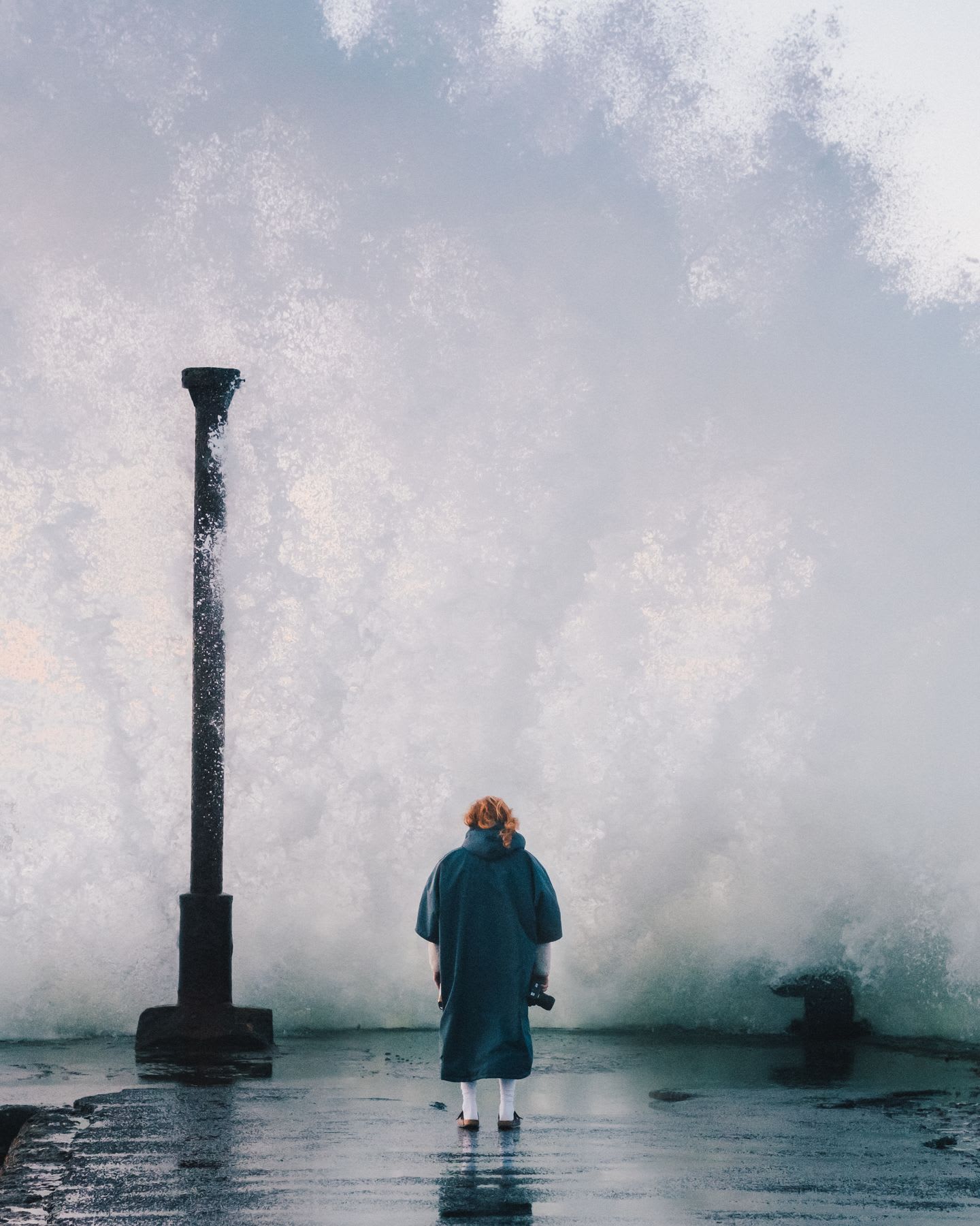 A woman in a blue raincoat stands on a wet pier facing a massive wave crashing into the structure, with mist and spray filling the air.