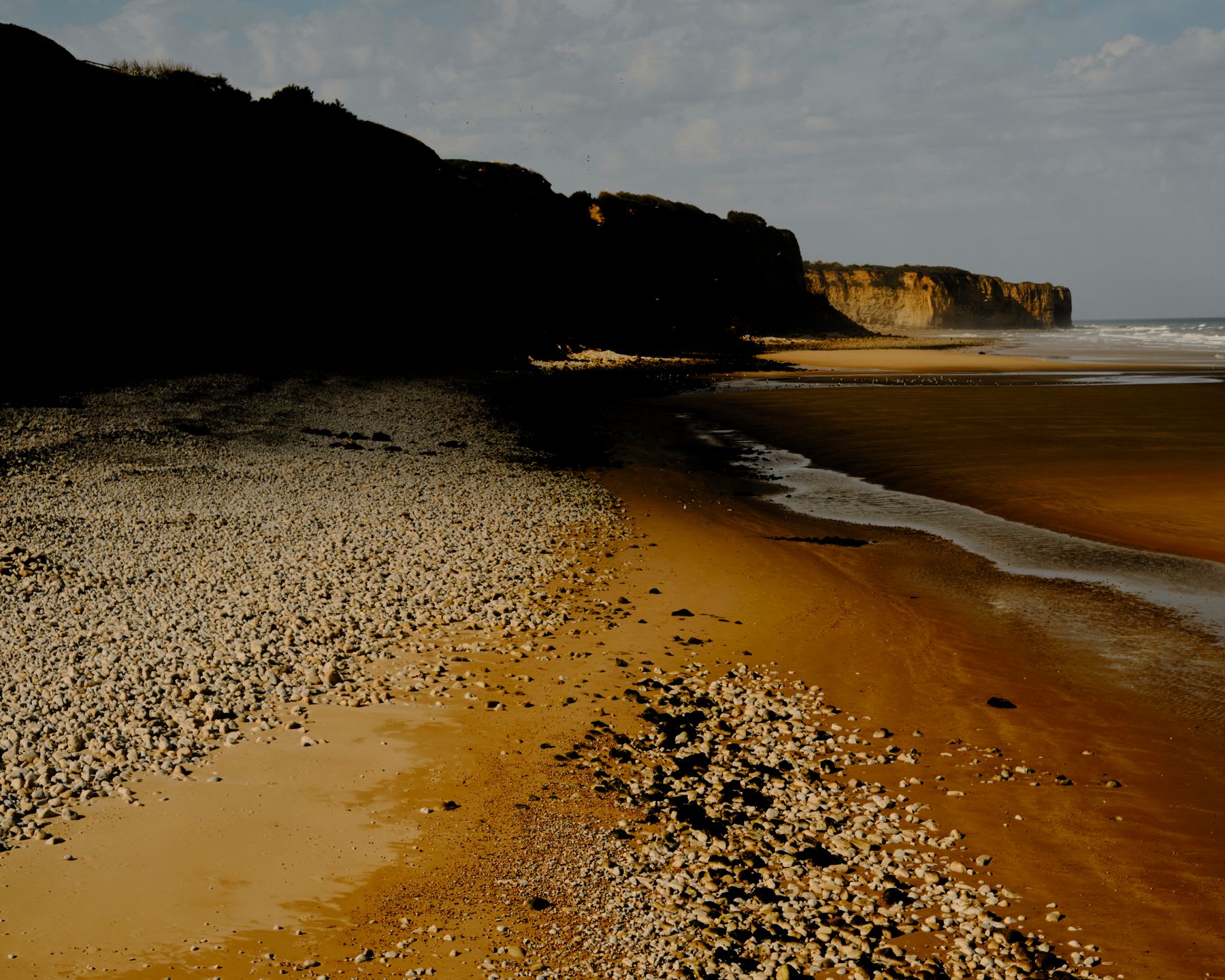 A rocky and sandy beach stretches alongside a dark cliff on the left, leading to an illuminated cliff face on the right, with ocean waves visible in the distance.