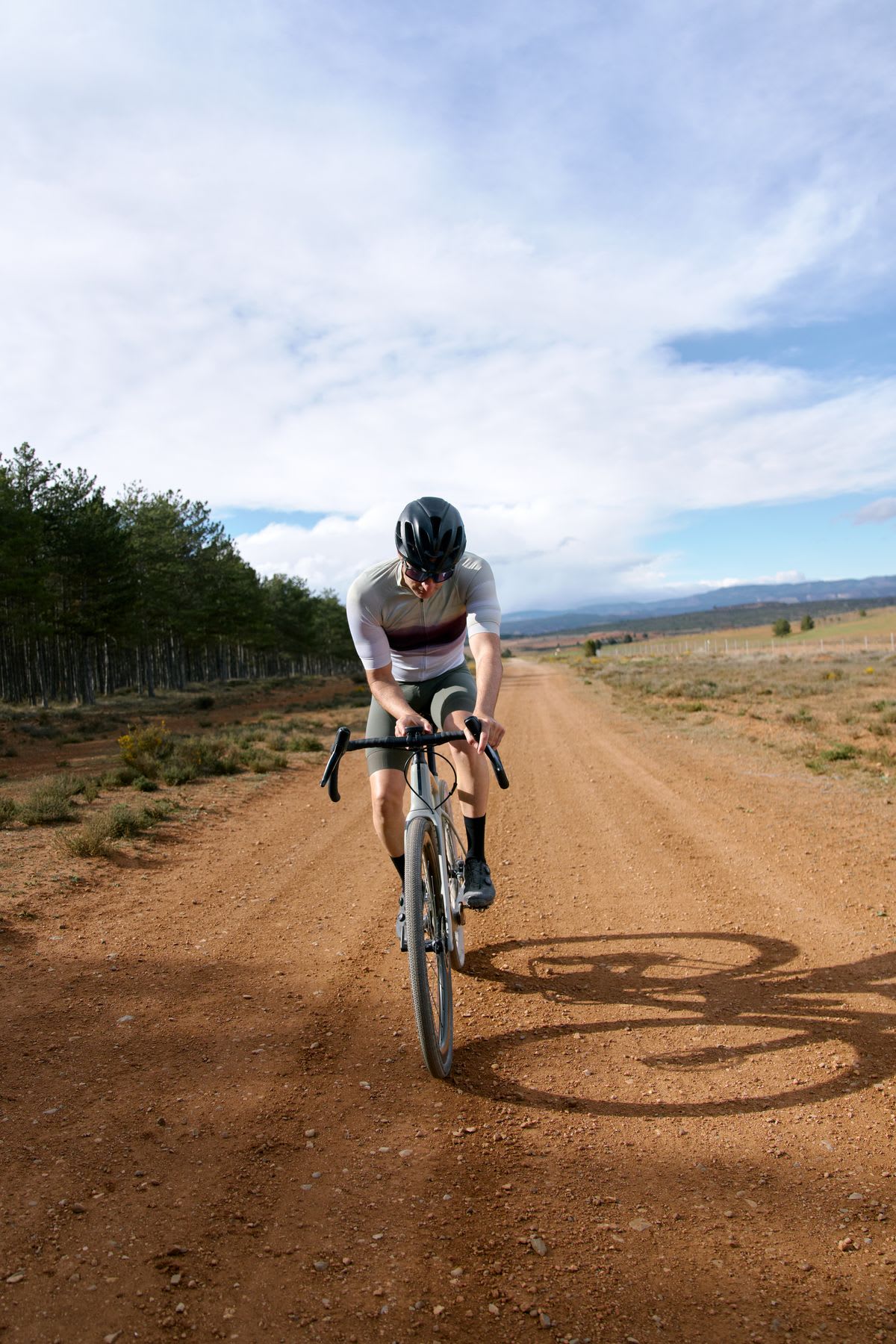 A male cyclist rides a bicycle on a dirt road with a forest on one side and a grassland on the other.