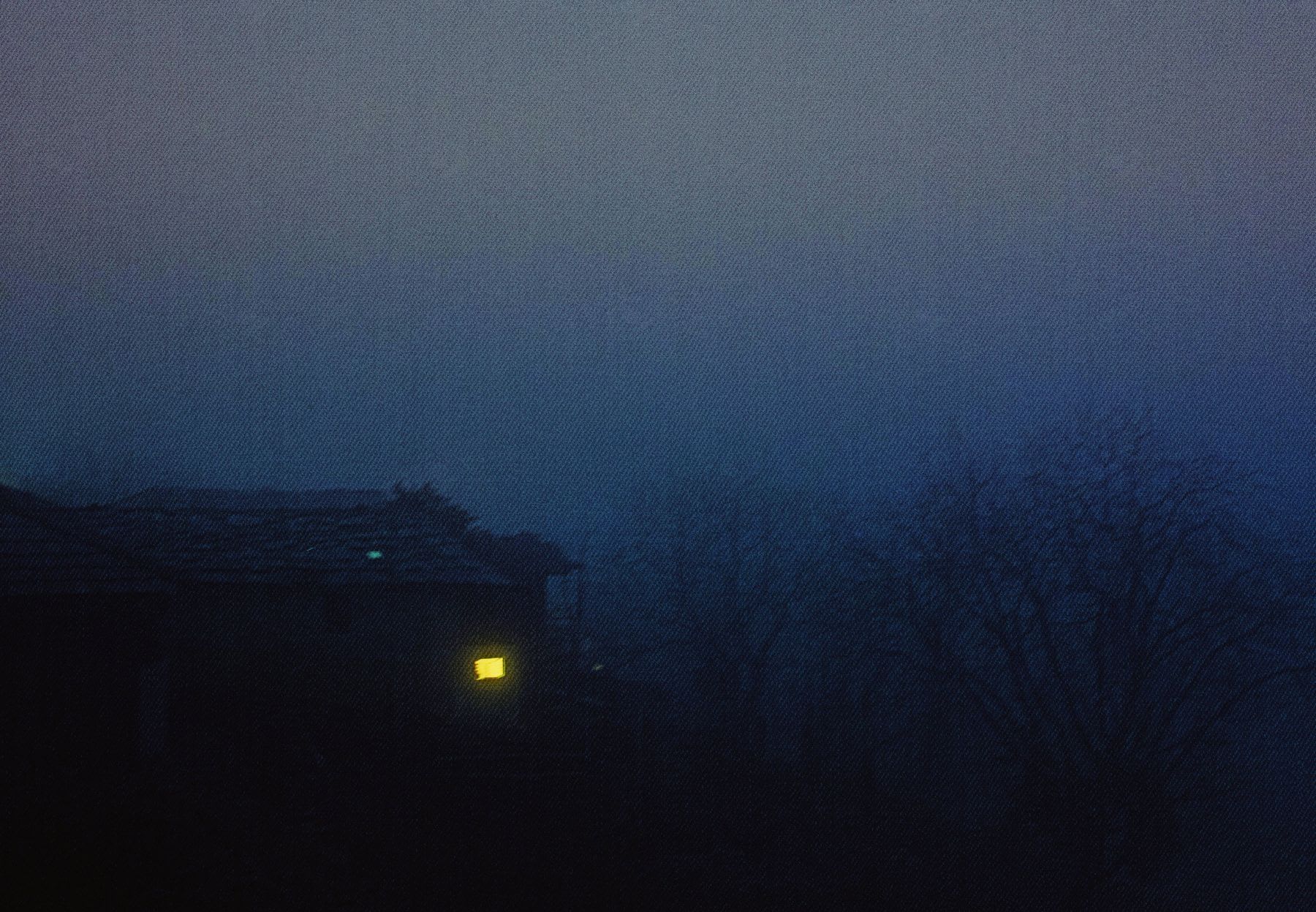 A golden light shines in the window of a rural house on a cloudy, winter evening.