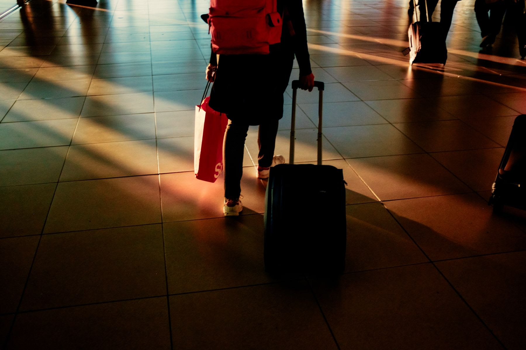 A traveler walks through an airport terminal pulling a suitcase with one hand and holding a shopping bag with the other, bathed in the warm light of the setting sun.