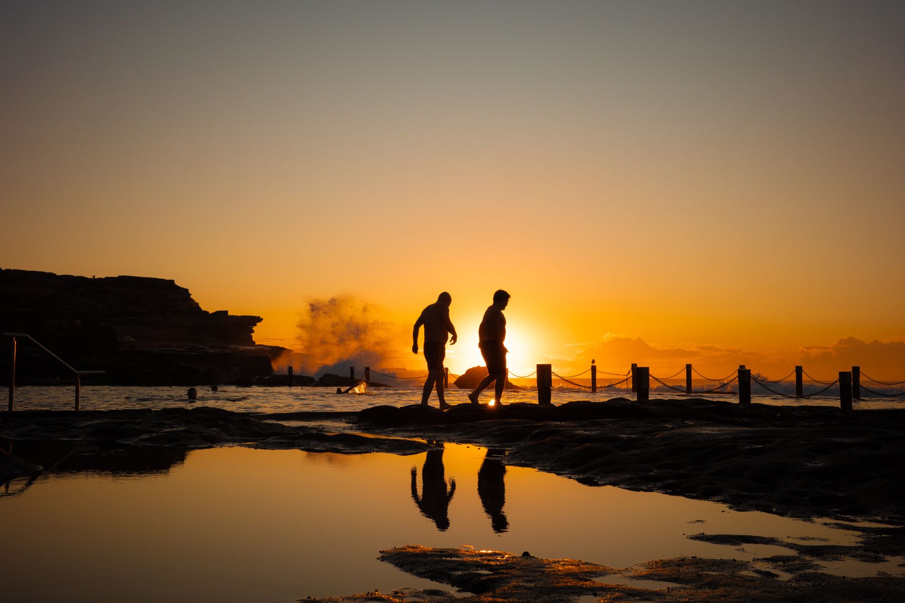 Two people are silhouetted against a golden sunrise as they walk along a rocky coast, their reflections cast in a tidal pool.