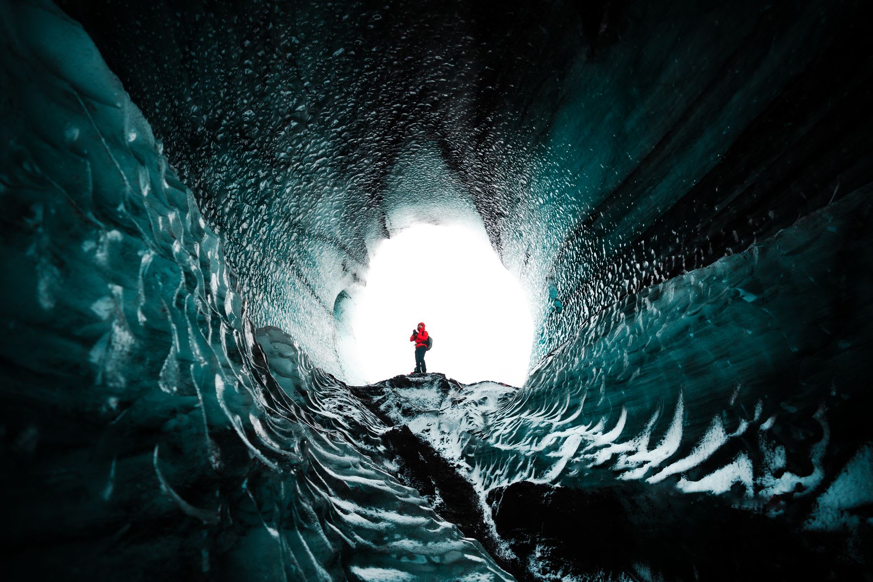 A person standing at the entrance of an ice cave, taking a picture of the inside.