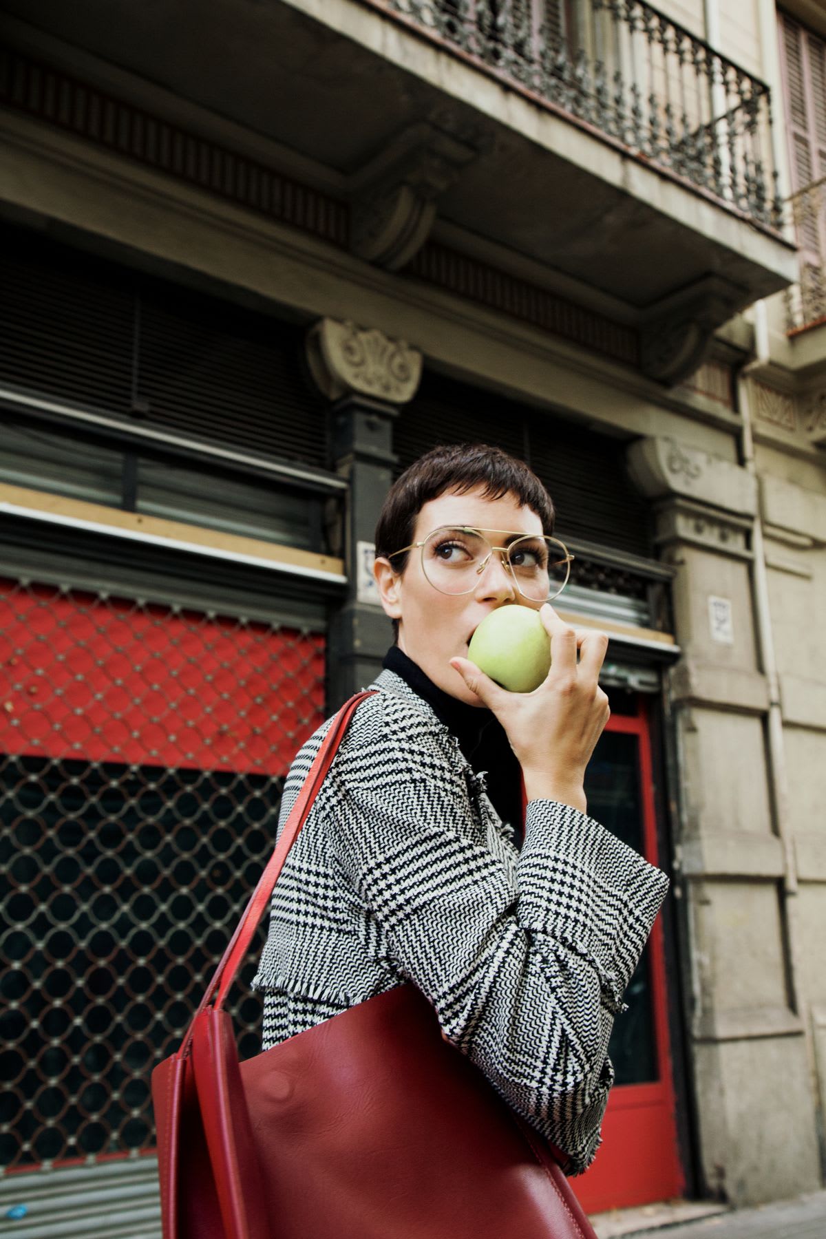 A stylish woman in a patterned coat and glasses eats a green apple while carrying a red handbag on a city street.