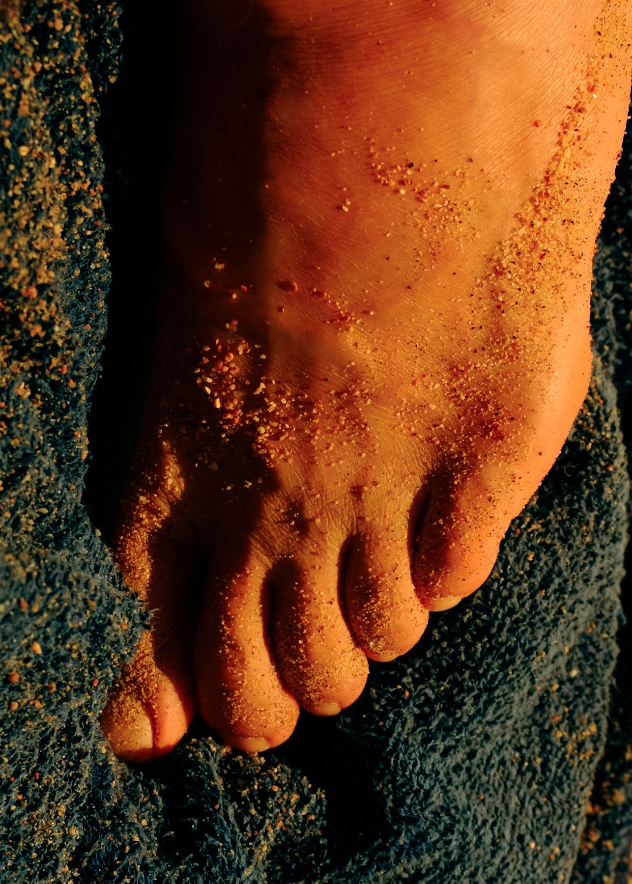 A close-up shot captures a person's sandy, bare foot resting on a blue towel in the warm glow of the sun.
