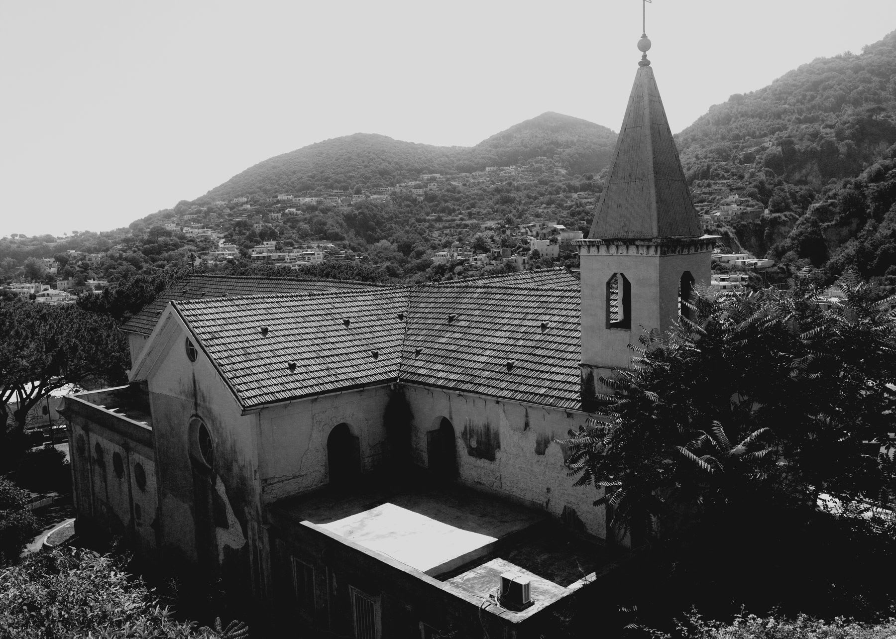 A black and white, high-angle view of an old monastery with a bell tower, nestled in a lush, hilly countryside village.