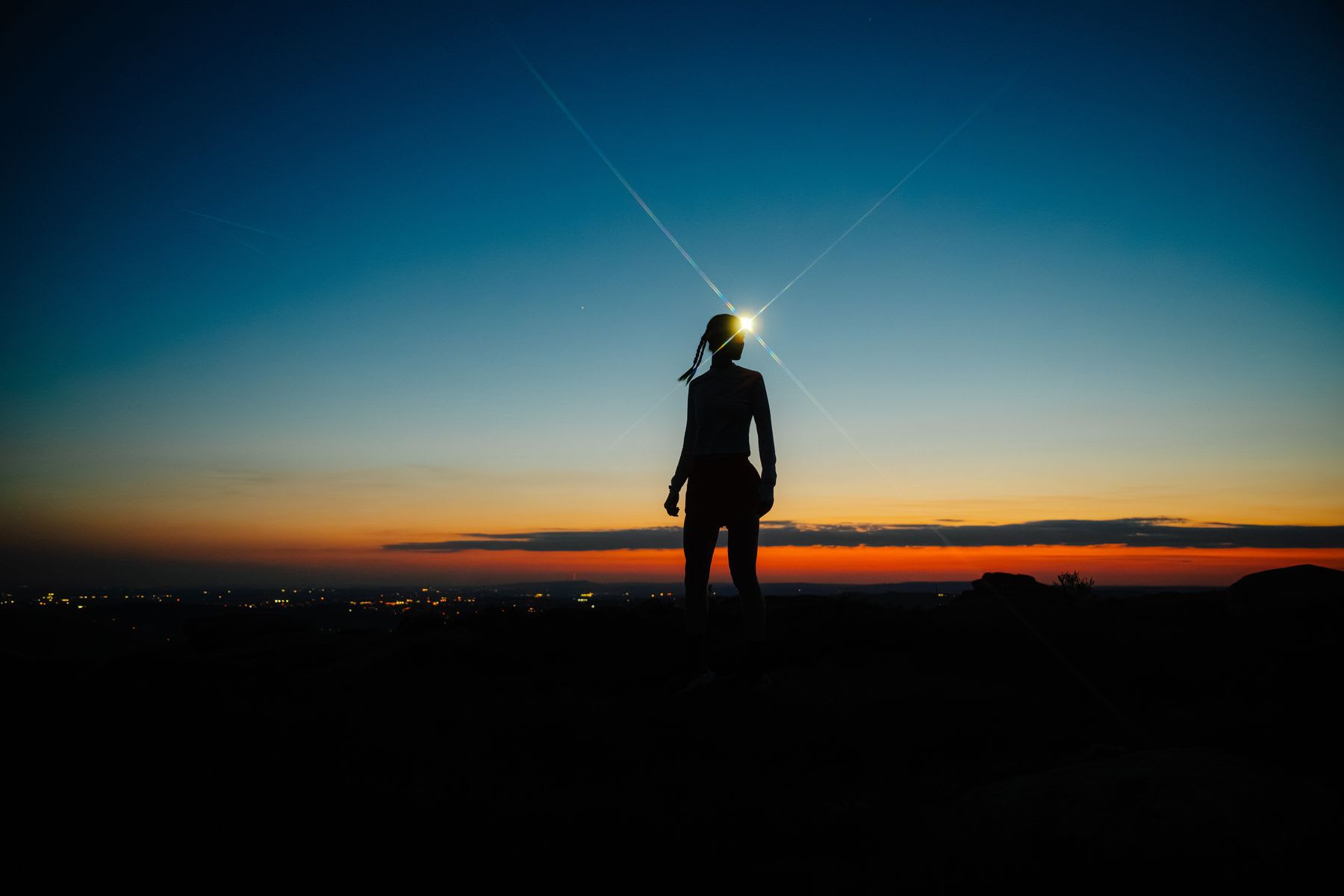 A woman stands in silhouette against a vibrant dusk sky, her headlamp shining brightly as she overlooks the city lights below.