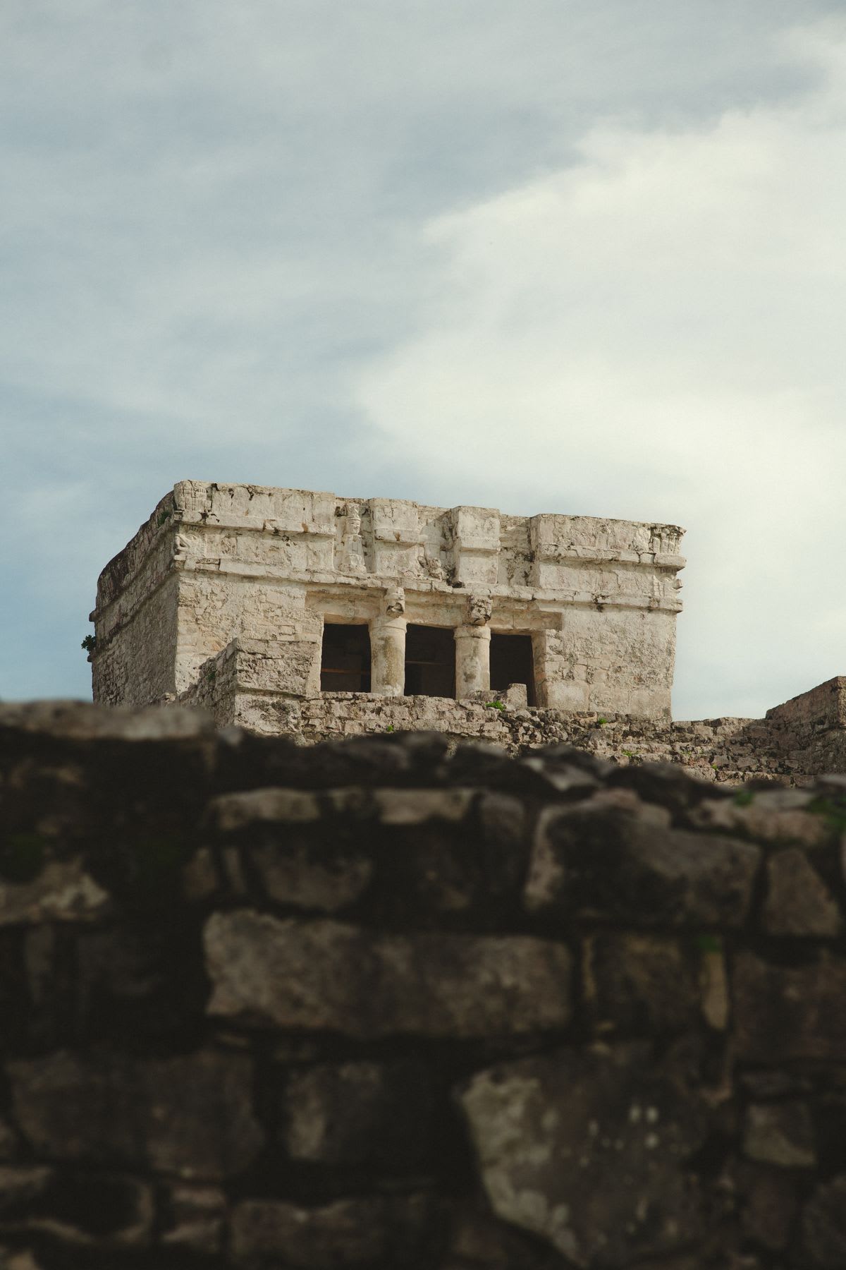 An ancient stone structure with columns is centered under a cloudy sky, with a blurry stone wall in the foreground.