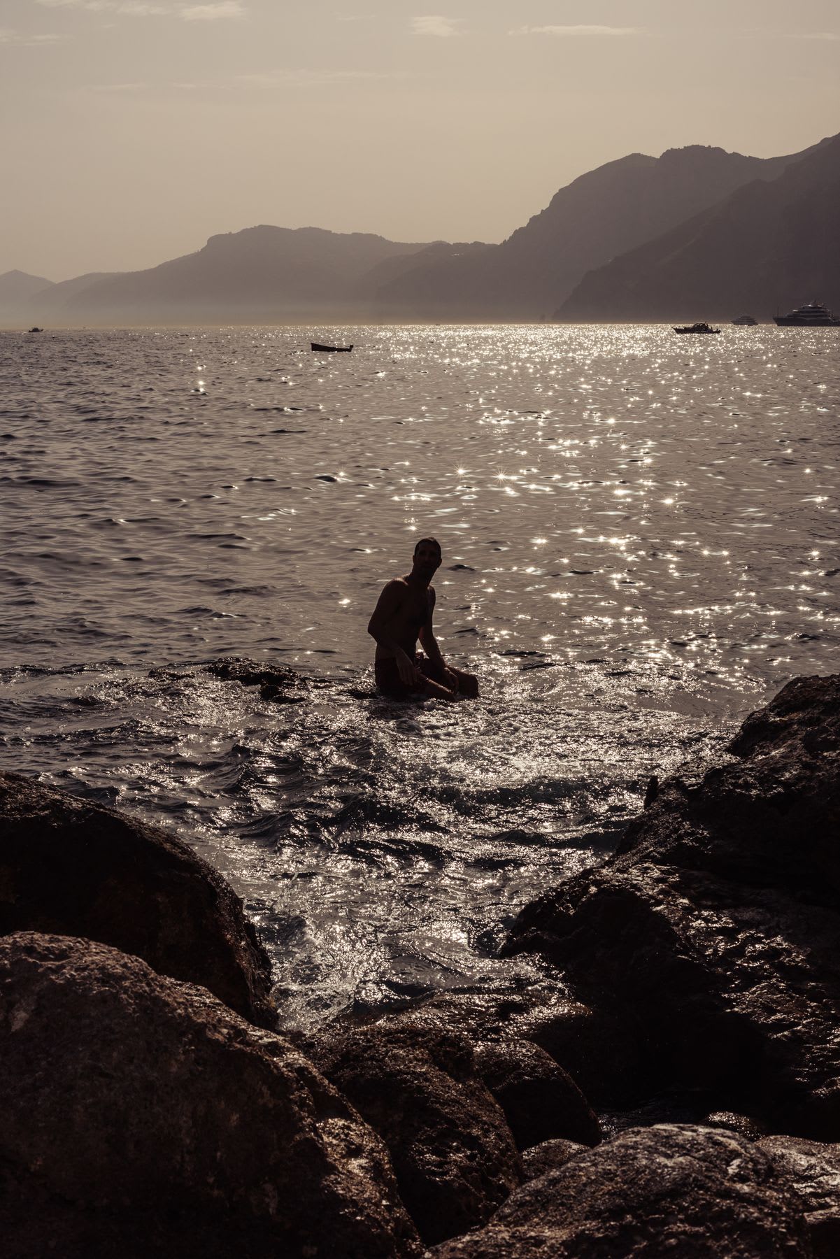 A silhouetted man sits on a rock in the water, with shimmering waves around him under a soft, hazy sky. In the background, distant mountains and boats are visible as the light gently reflects off the water.