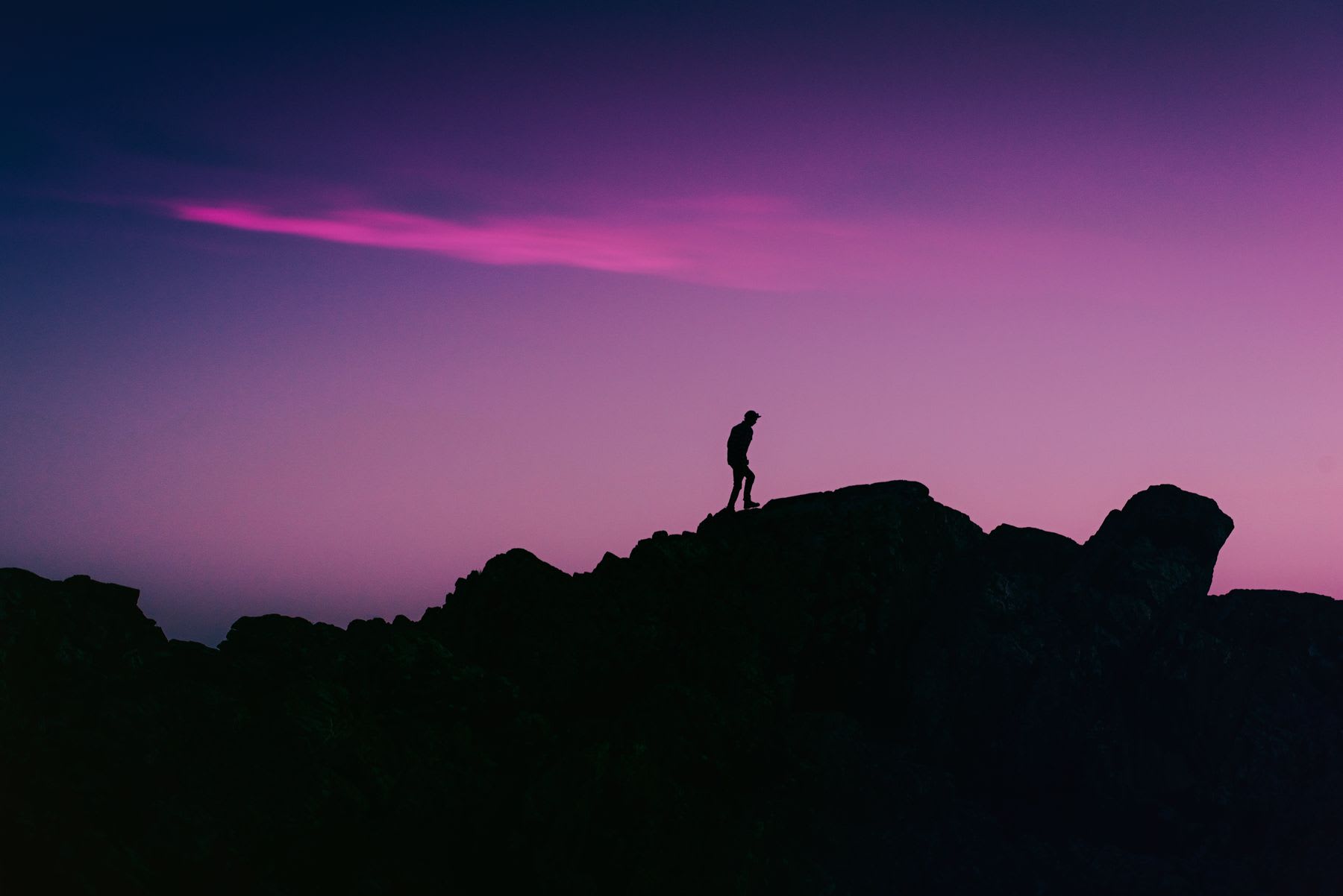 A lone hiker is silhouetted against a stunning purple and pink sky while standing on a rocky mountain ridge at dusk.