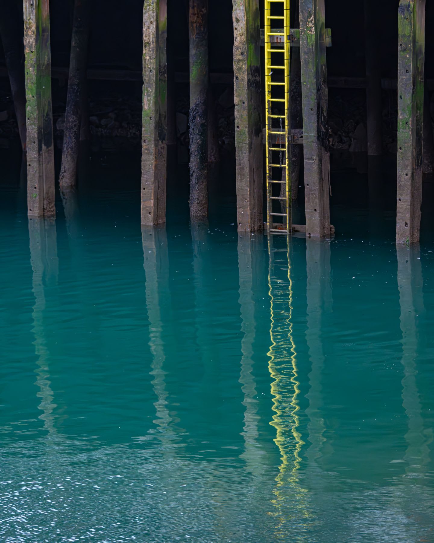 A yellow staircase attached to vertical wooden pillars on a pier, with turquoise water all around.