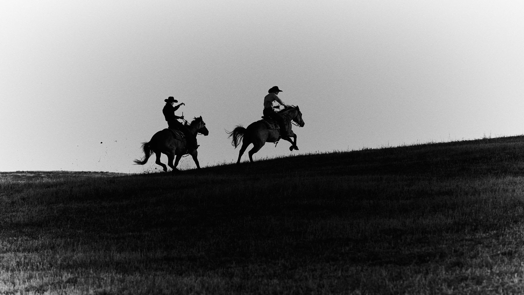 Two horse riders ride over a hill quickly during the day.