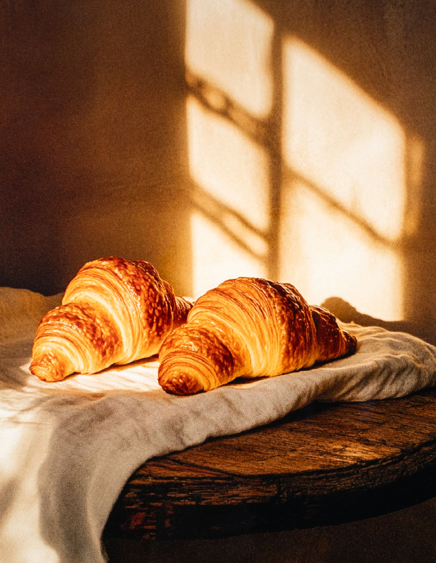 Two croissants on a cloth as sunlight from a window falls on them.