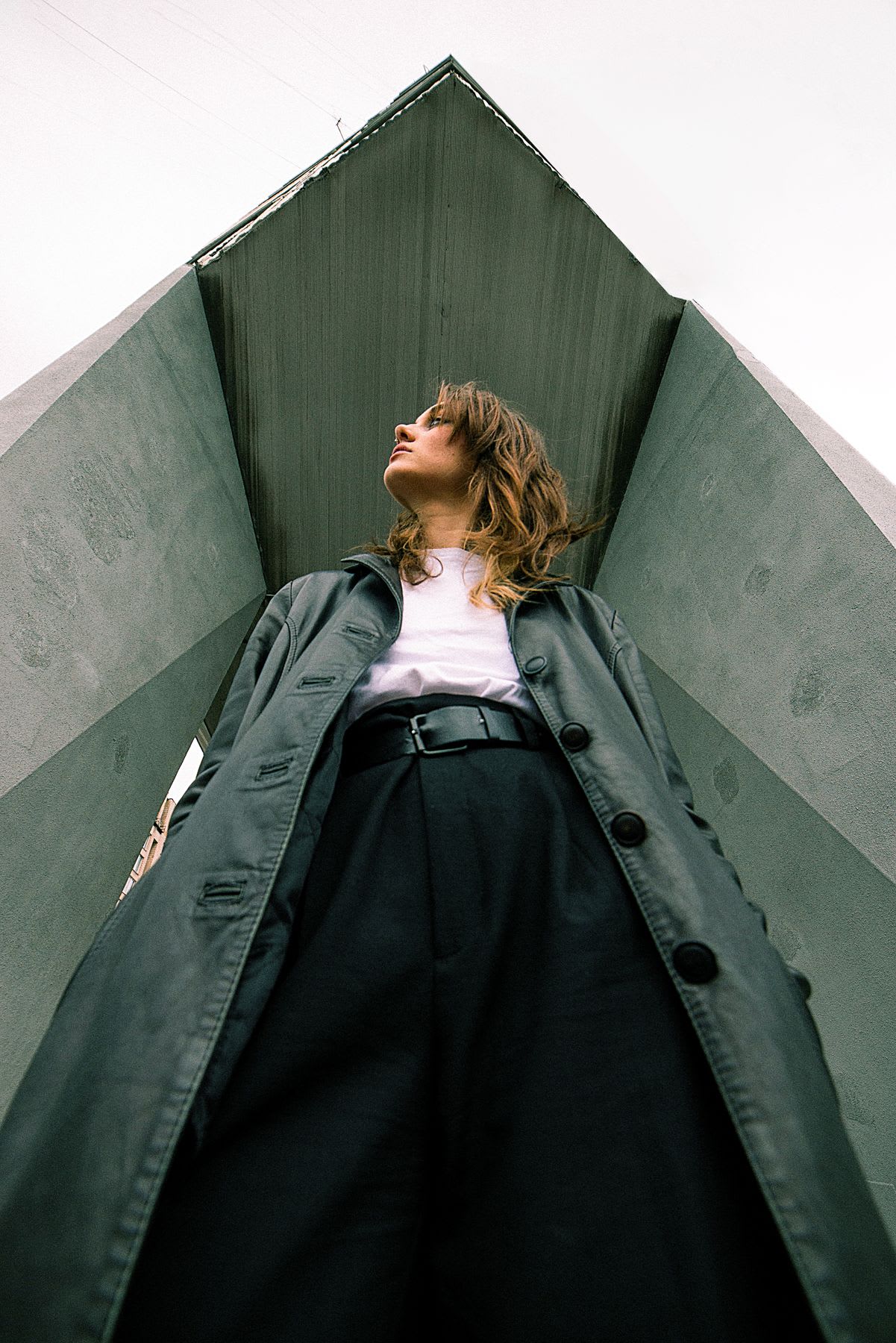 A low-angle shot captures a stylish woman in a dark trench coat looking up while standing between imposing concrete structures.