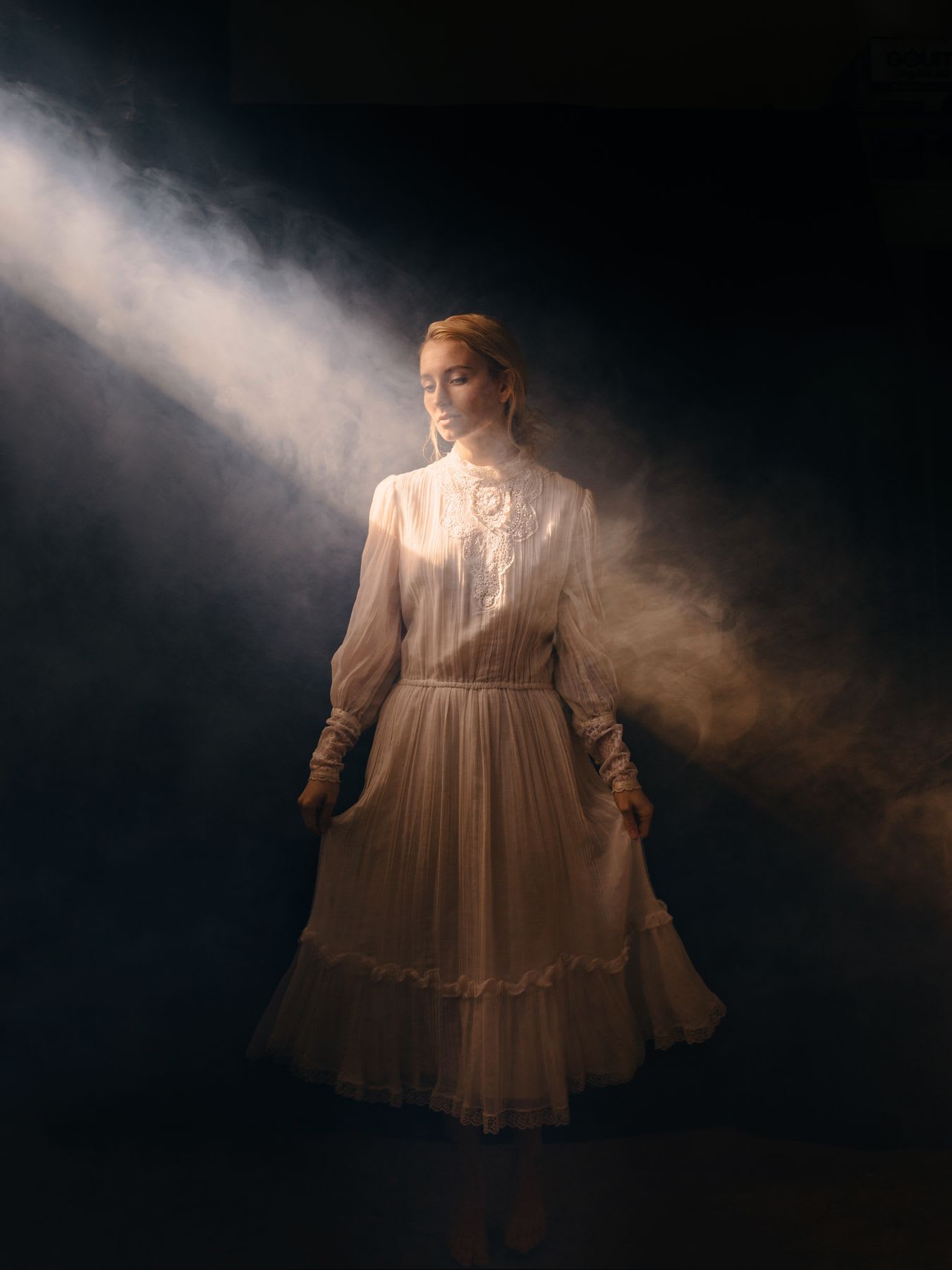 Woman in a vintage lace dress standing in a beam of light with a hazy smoke effect around her.