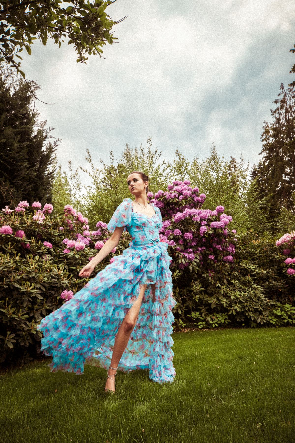 a woman posing gracefully in a voluminous, flowing blue and pink floral dress with ruffles and a high slit, standing on a lush green lawn surrounded by vibrant pink rhododendron bushes and tall trees under an overcast sky.