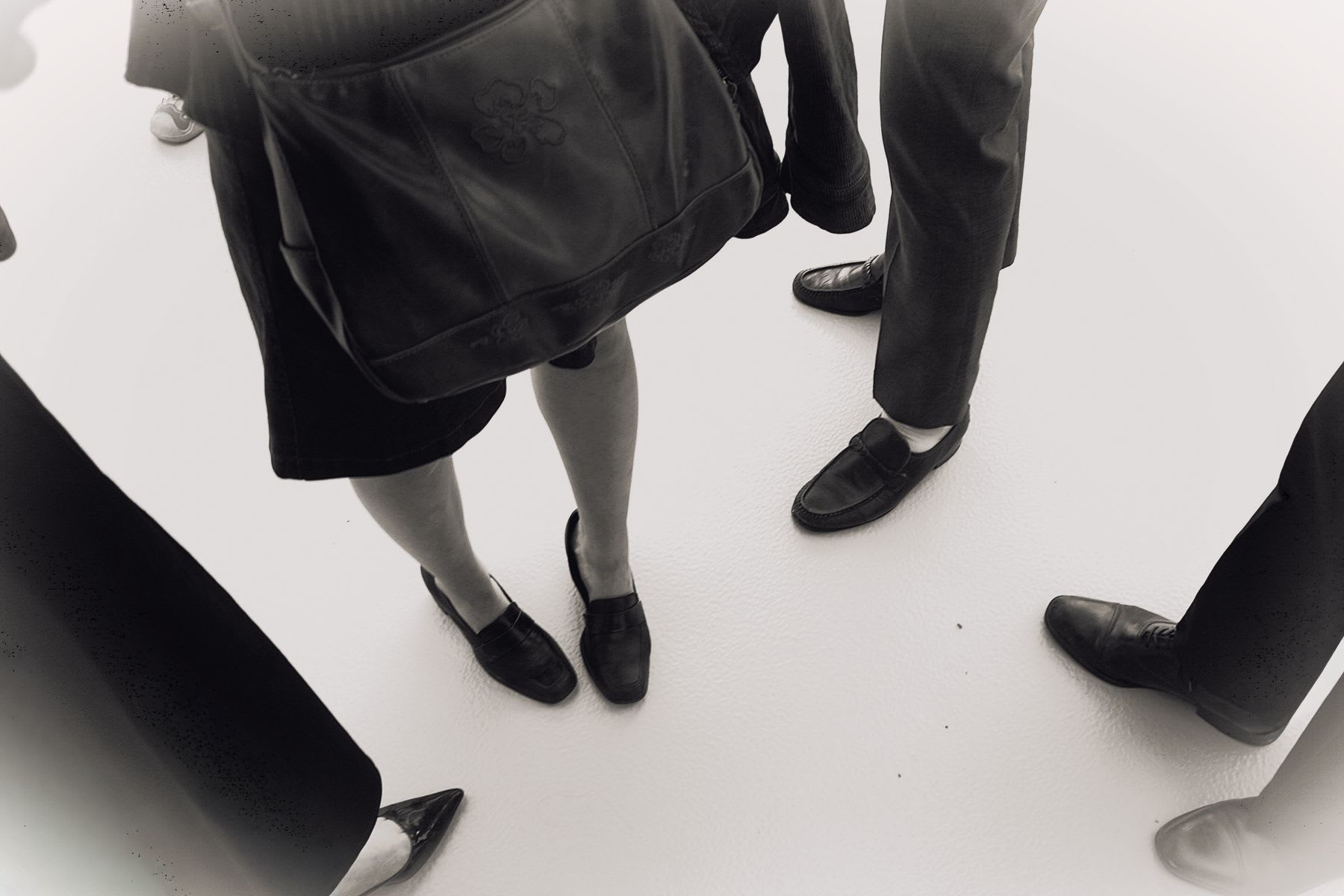 A high-angle, black and white view of several people in formal wear standing together, focusing on their shoes and a handbag.
