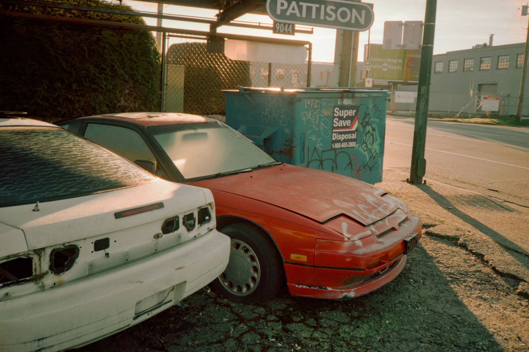 Two damaged and weathered sports cars, one red and one white, are parked in a gritty urban lot next to a graffiti-covered dumpster.
