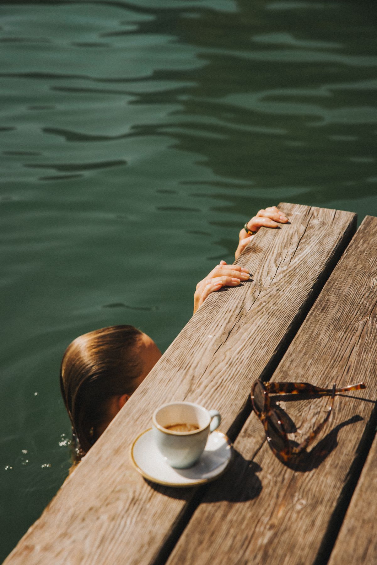 A woman with wet hair holds onto a wooden dock while swimming, with a cup of coffee and sunglasses resting on the sunlit planks.