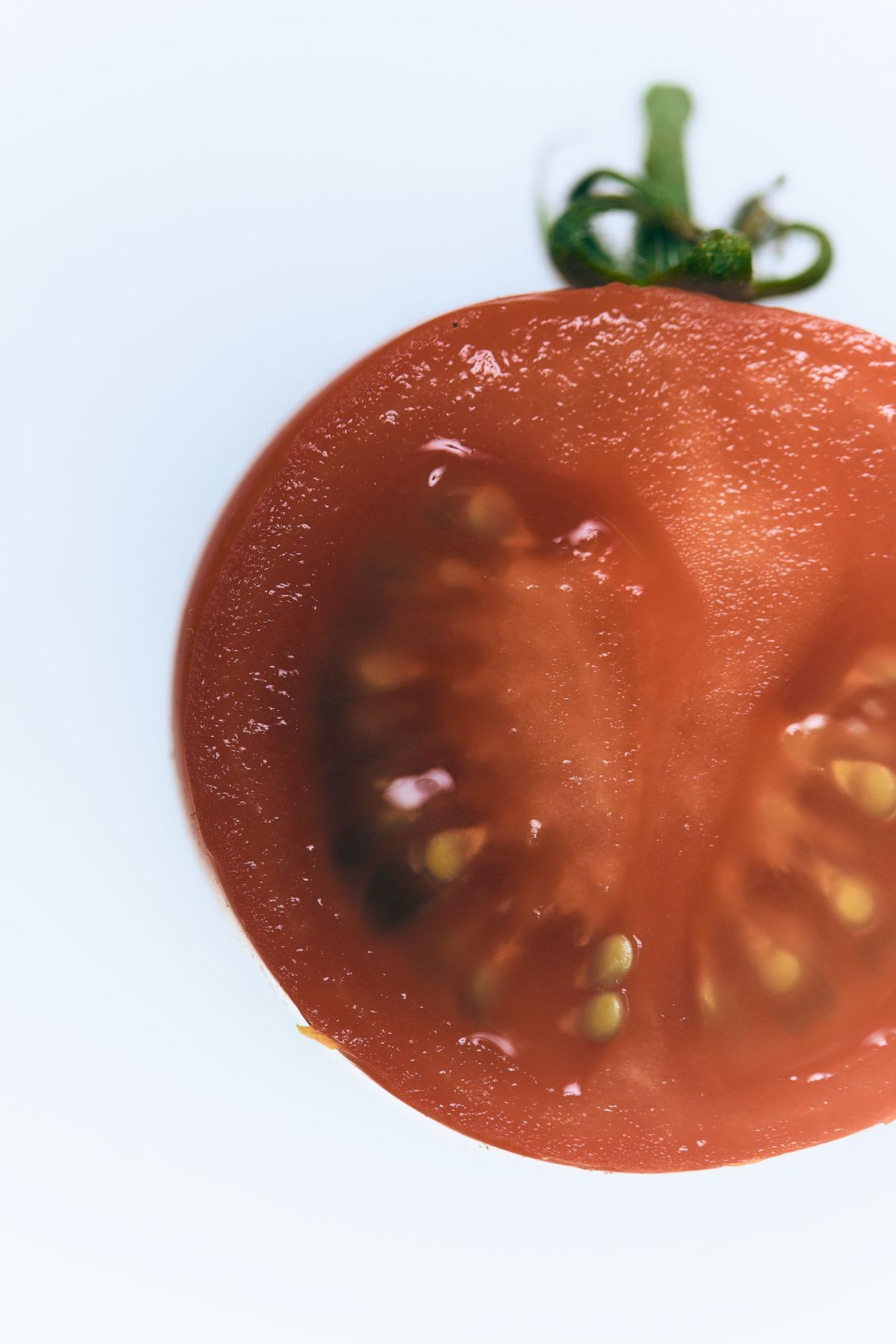 An extreme close-up shot of a freshly sliced red cherry tomato with its green stem, showcasing the juicy pulp and seeds against a white background.