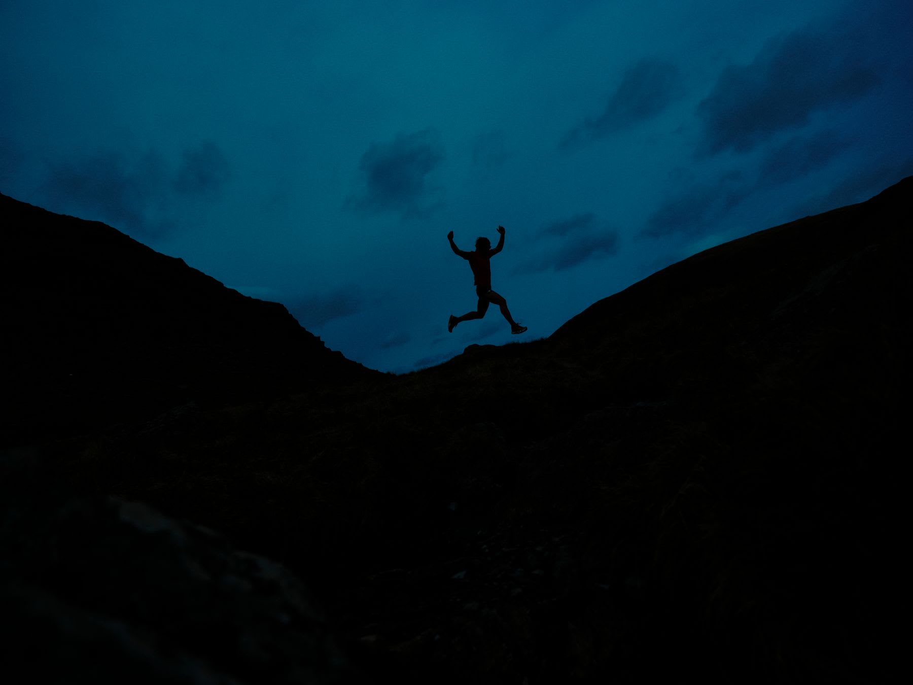 A person is silhouetted jumping with arms raised in celebration between two dark hills against a moody blue sky at night.