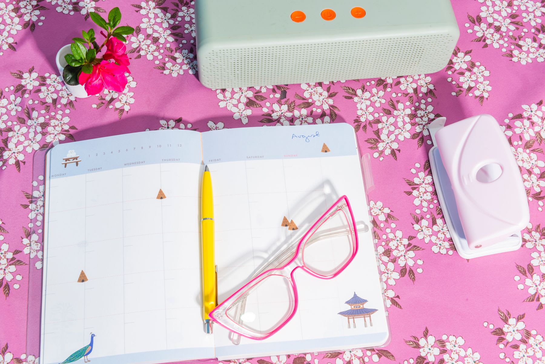 Open planner on a floral pink tablecloth with a yellow pen and pink glasses next to a potted plant, a portable speaker, and a pink leaf hole punch.