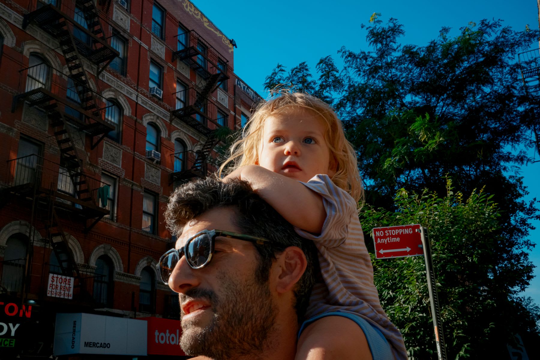 A little girl with blonde hair and blue eyes sits on her father's shoulders as they walk through a city on a sunny day.