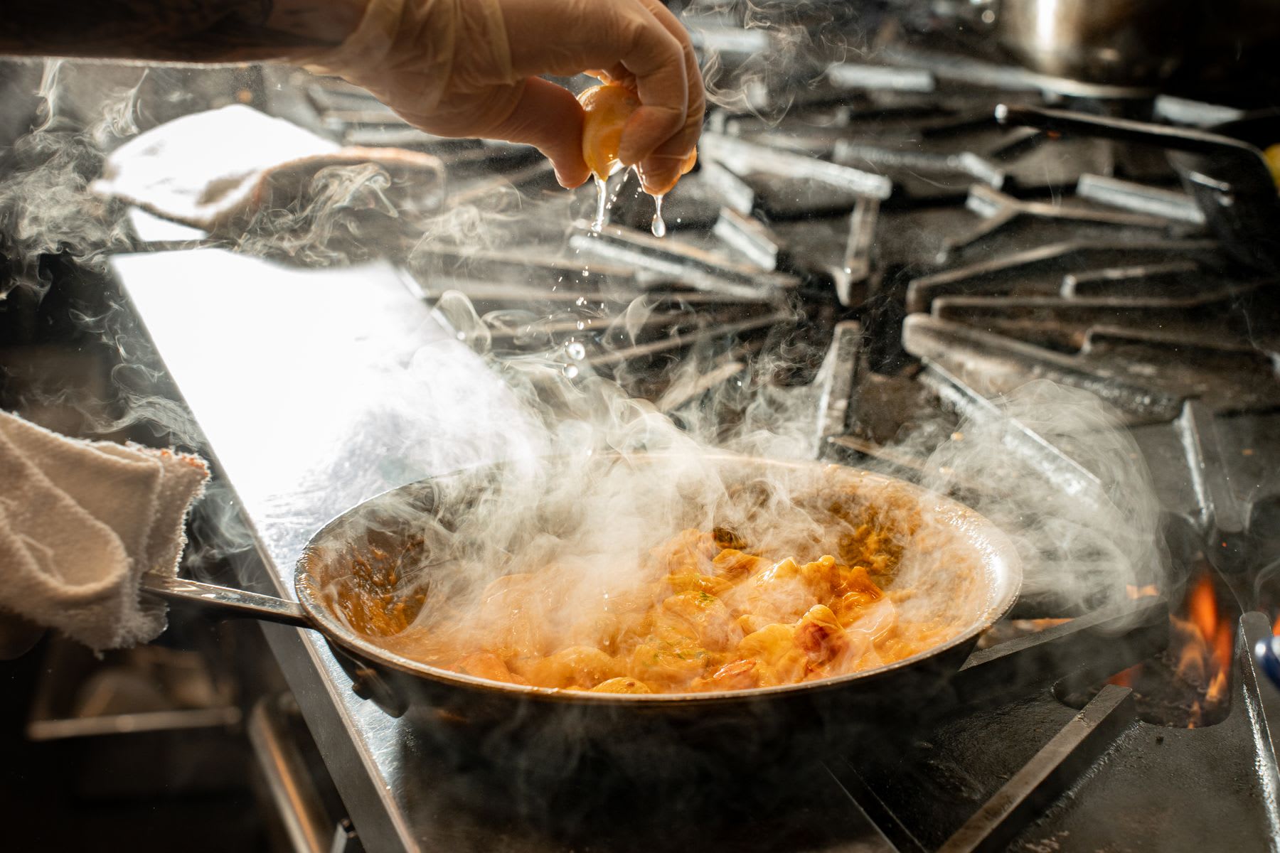A person squeezing a lemon wedge over a steaming skillet of cooking food.