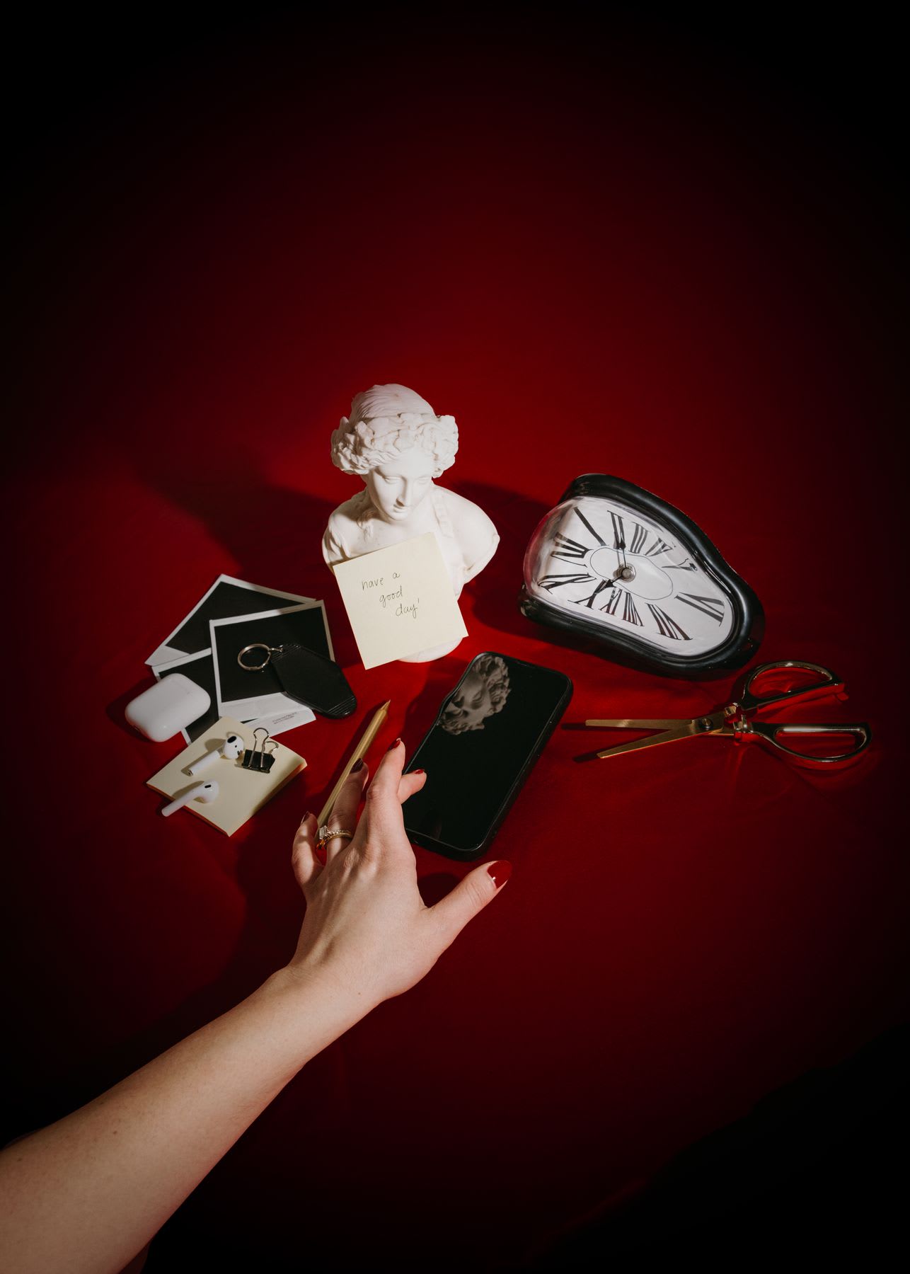 Hand reaching for a black smartphone on a red surface surrounded by various objects including a white bust sculpture with a note that says 'have a great day,' a melting clock, a pair of gold scissors, AirPods, Polaroid photos, and a keychain.