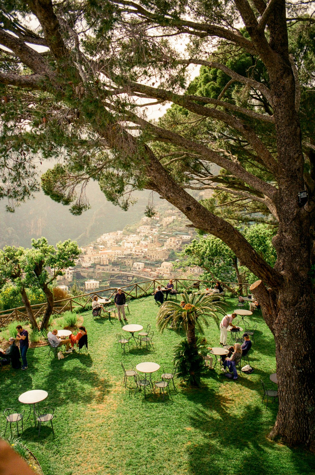A serene, tree-shaded garden terrace overlooks a picturesque hillside town, with people relaxing at scattered tables amidst lush greenery.