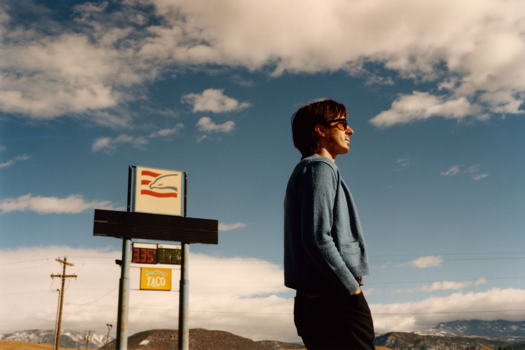 Man stands outdoors with a commercial sign and mountains in the background on a cloudy day.