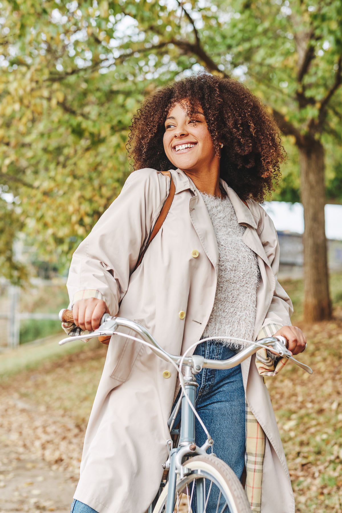 A happy woman with voluminous curly hair smiles brightly while holding her bicycle in a park during autumn.