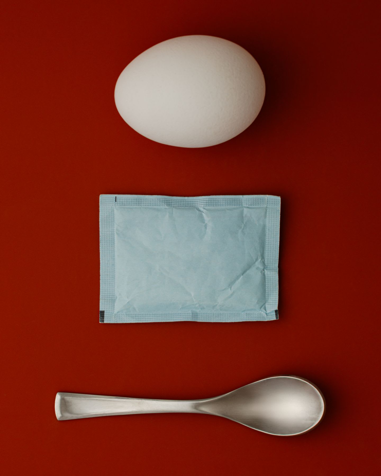 A white egg, a light blue packet, and a silver spoon are arranged vertically on a solid red background.